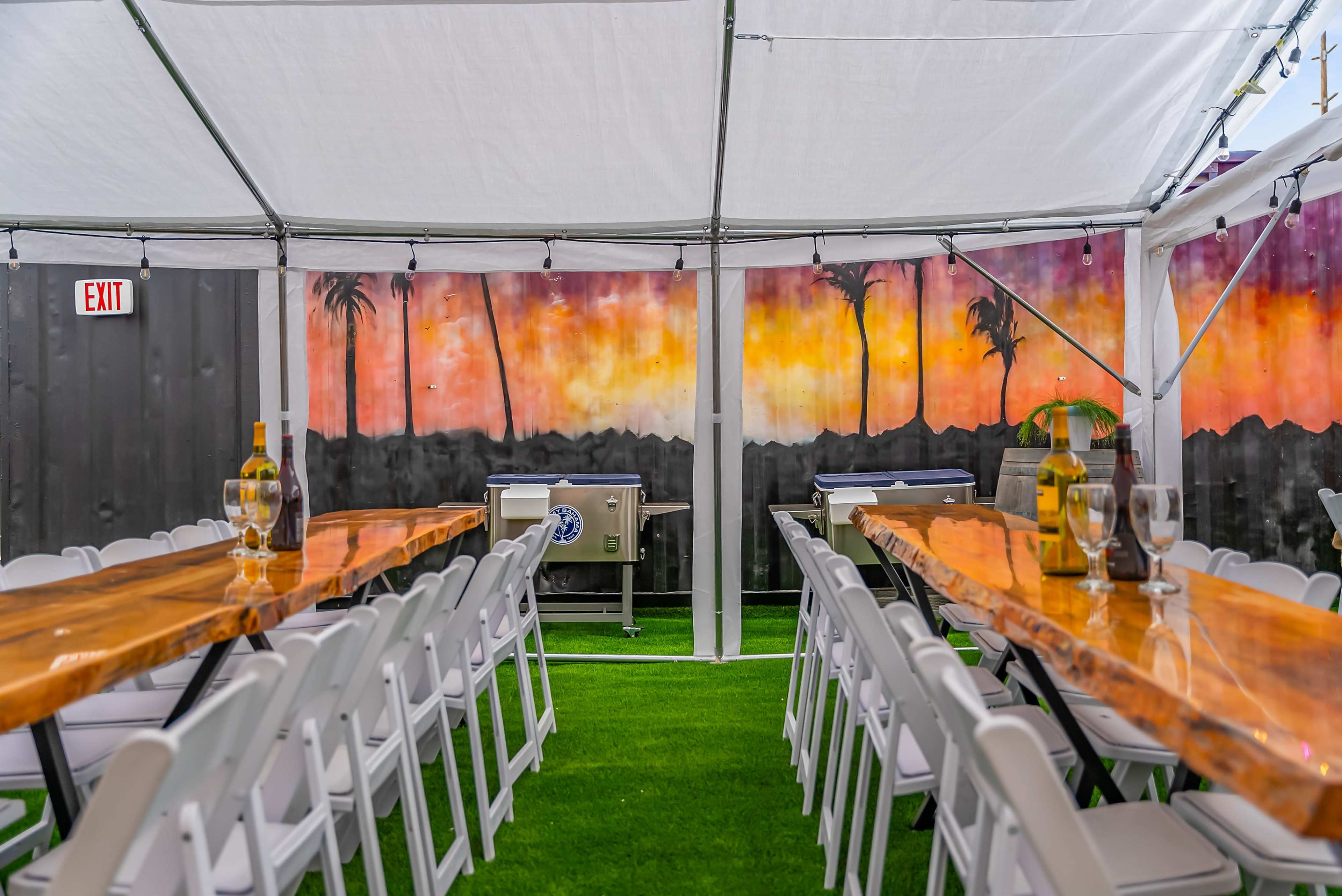 The image shows a tented dining area with long wooden tables arranged in rows, surrounded by a painted mural of a sunset and palm trees on the back wall.