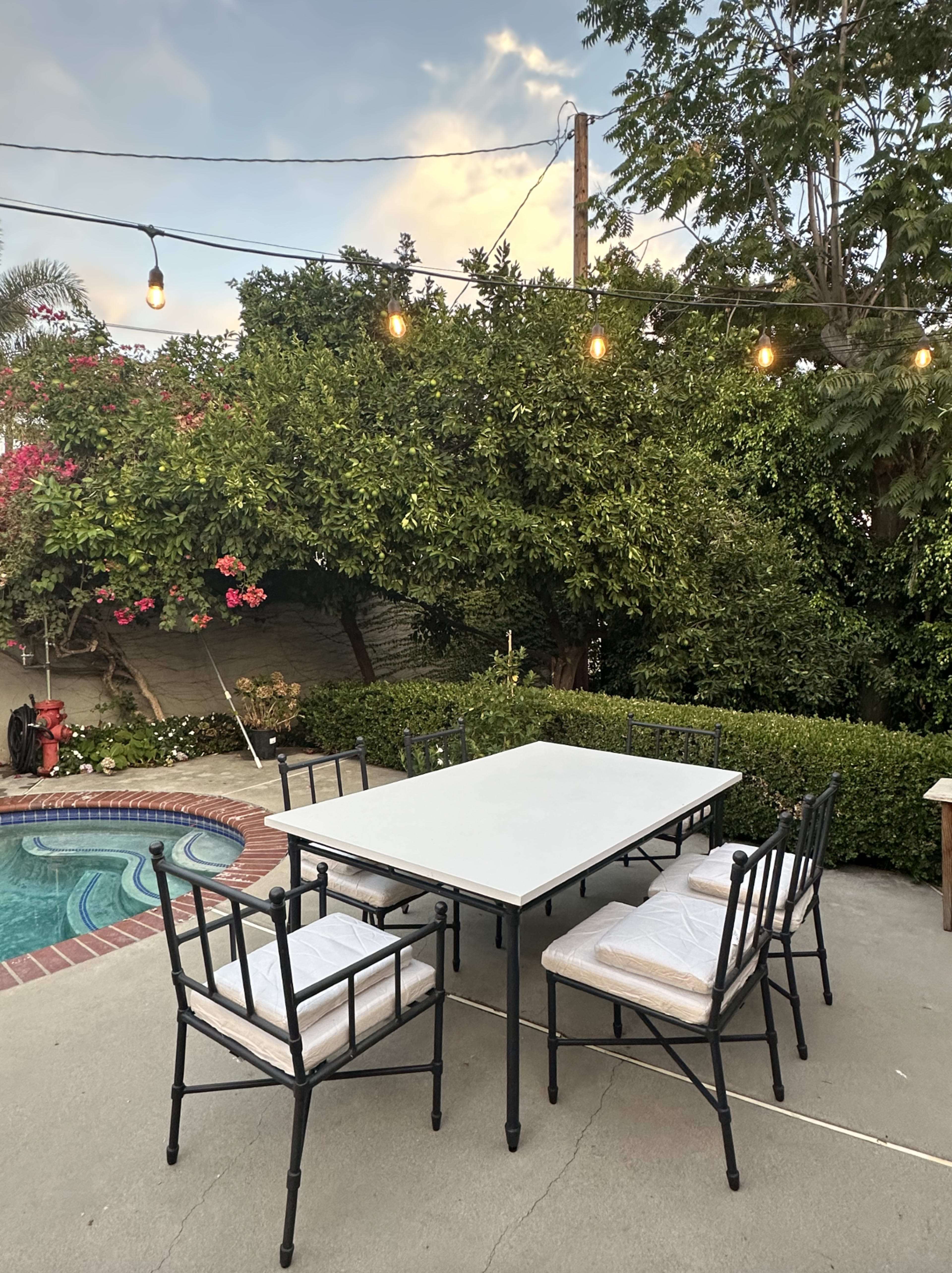 A patio area features a rectangular table with six chairs, surrounded by lush greenery and outdoor string lights above a nearby pool.