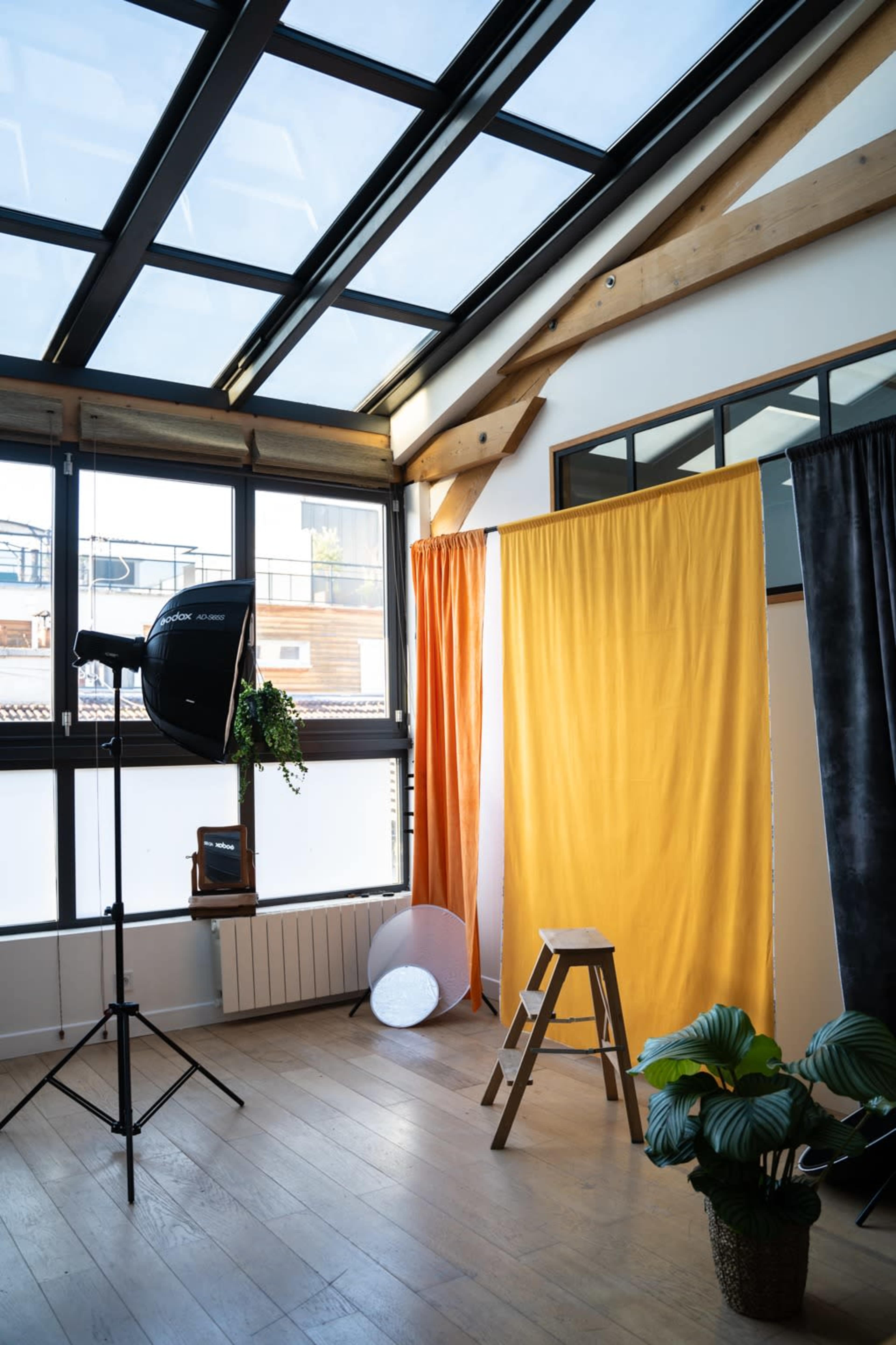 A well-lit studio space with a wooden floor, a photography backdrop with orange and yellow curtains, a stool, a light stand, and a potted plant.