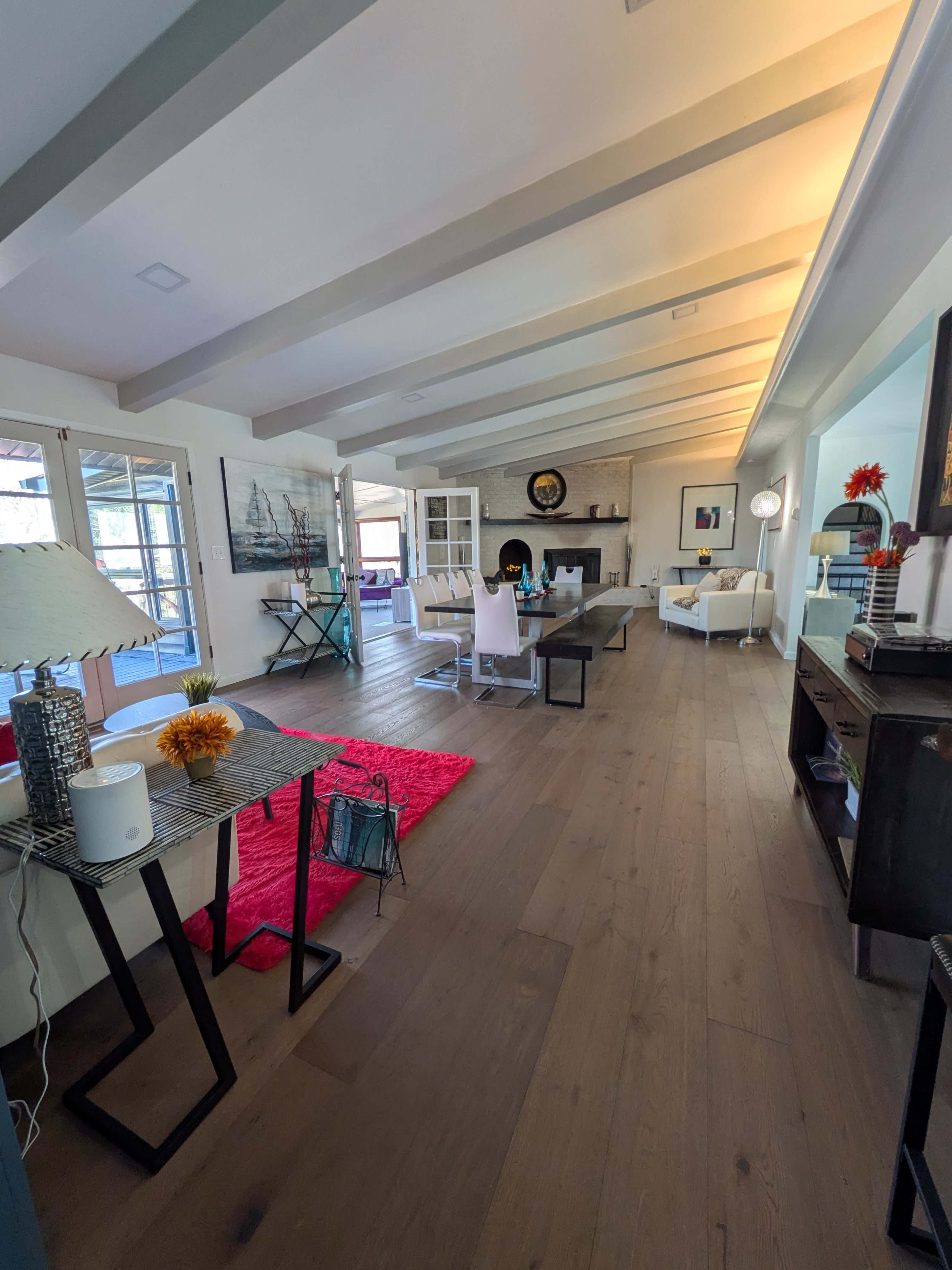 The image shows a spacious and modern living area with wooden flooring, a red area rug, and large windows that allow natural light to flood the room.