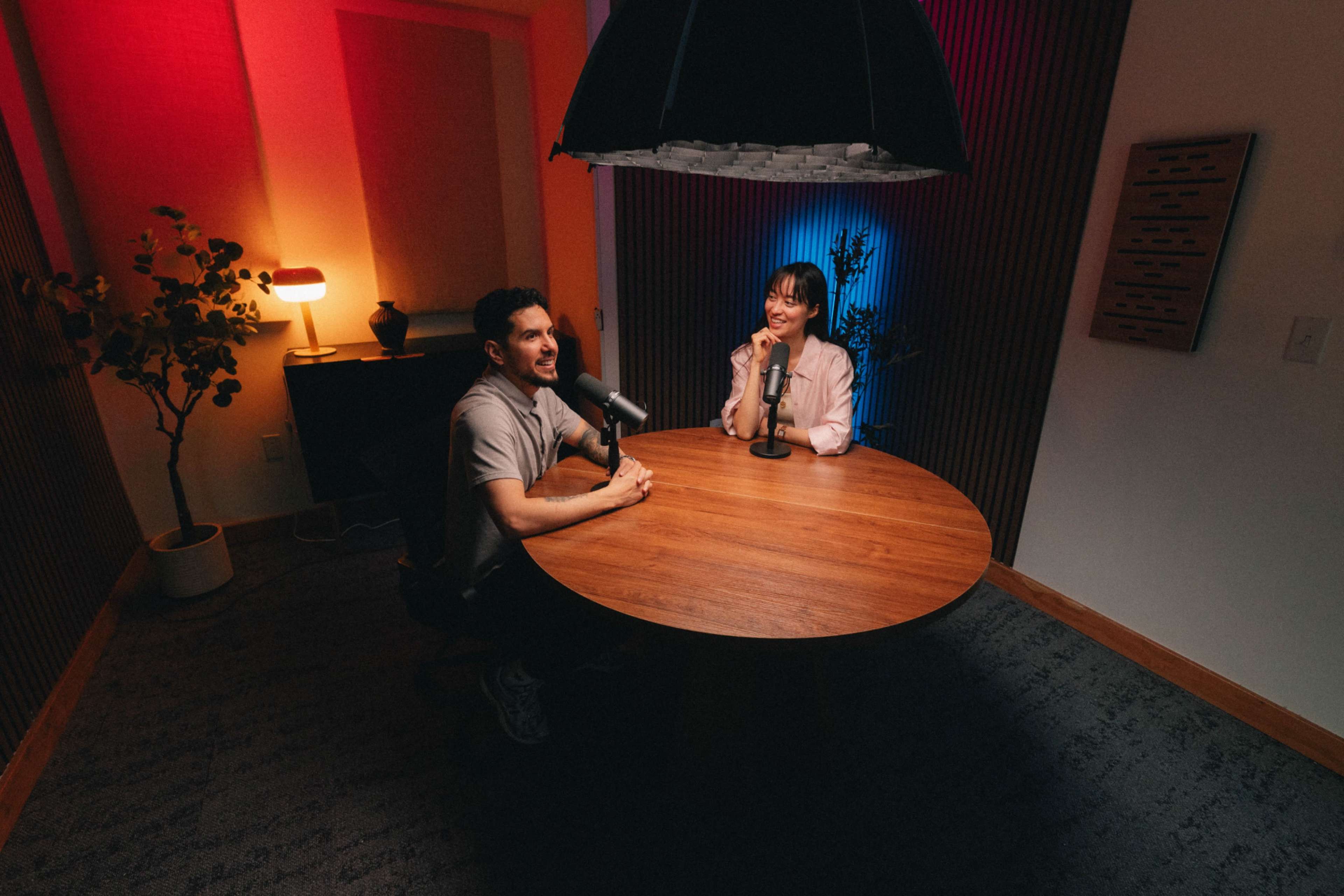 A man and a woman are seated at a round wooden table with microphones in a softly lit room designed for podcasting.