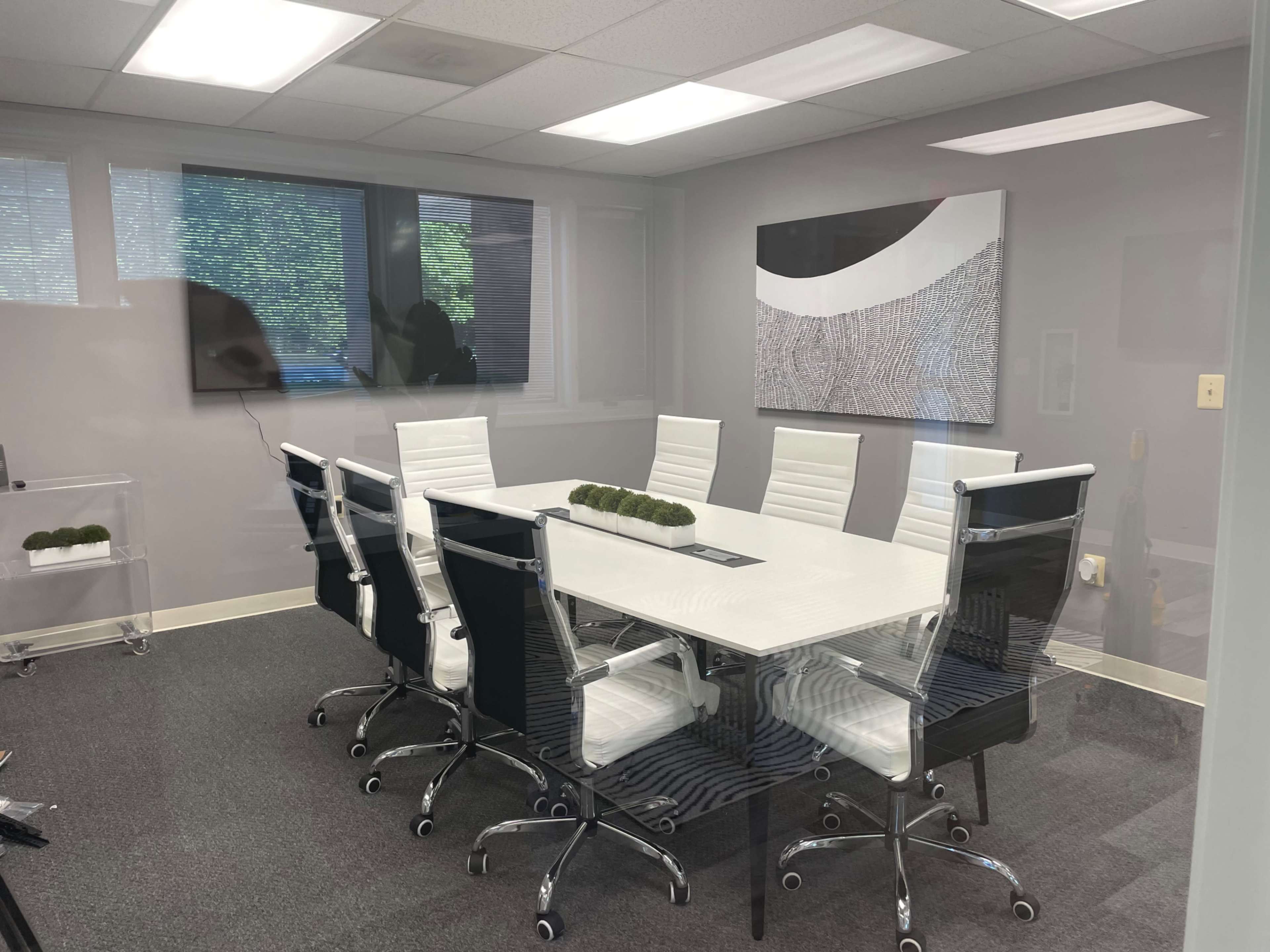 A modern conference room with a rectangular white table surrounded by black and white chairs, and two large screens mounted on the walls.