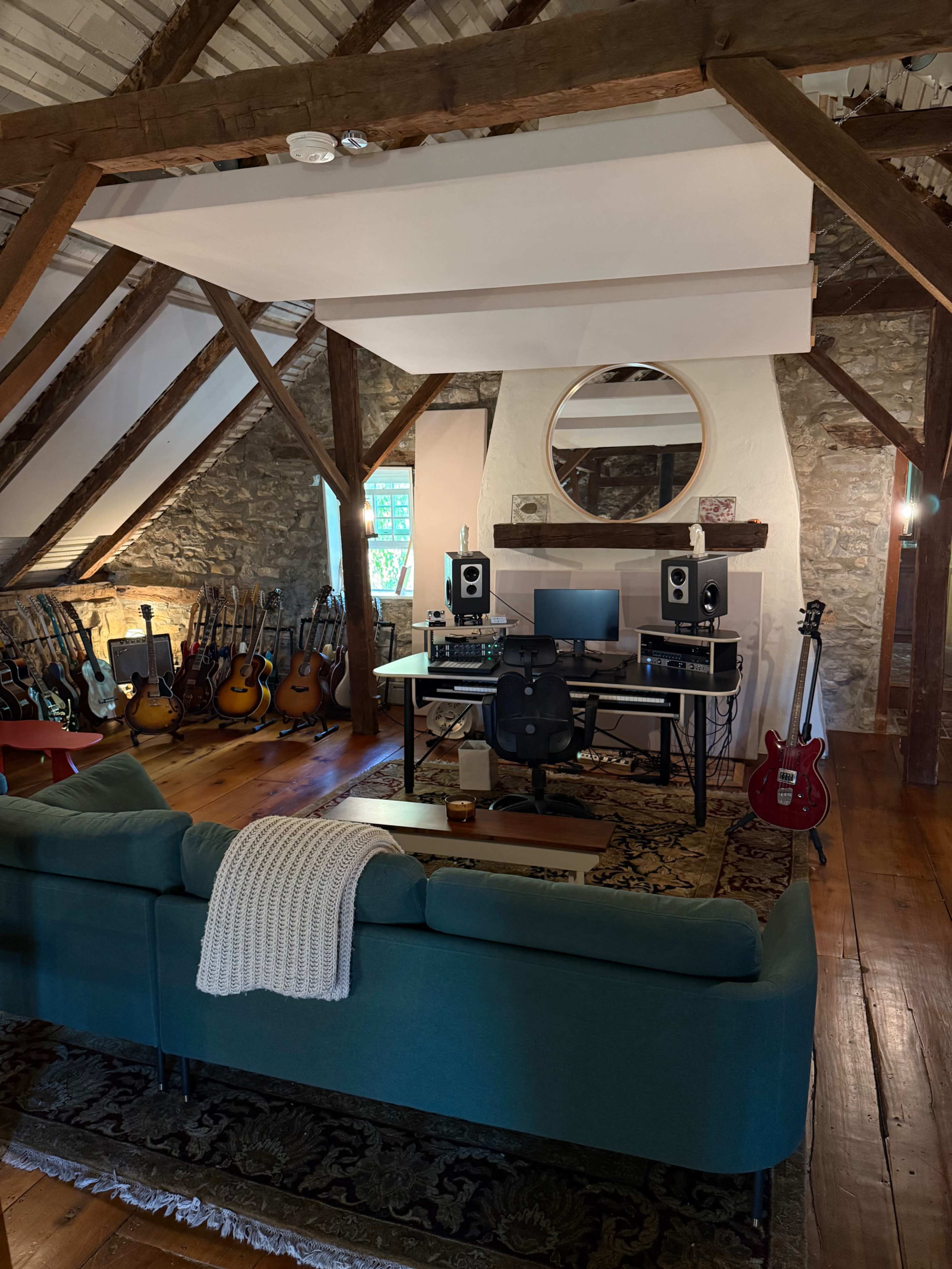 The image shows a music studio in an attic with wooden beams, a large circular mirror, a desk with computer equipment, a black office chair, and various guitars displayed on the walls.
