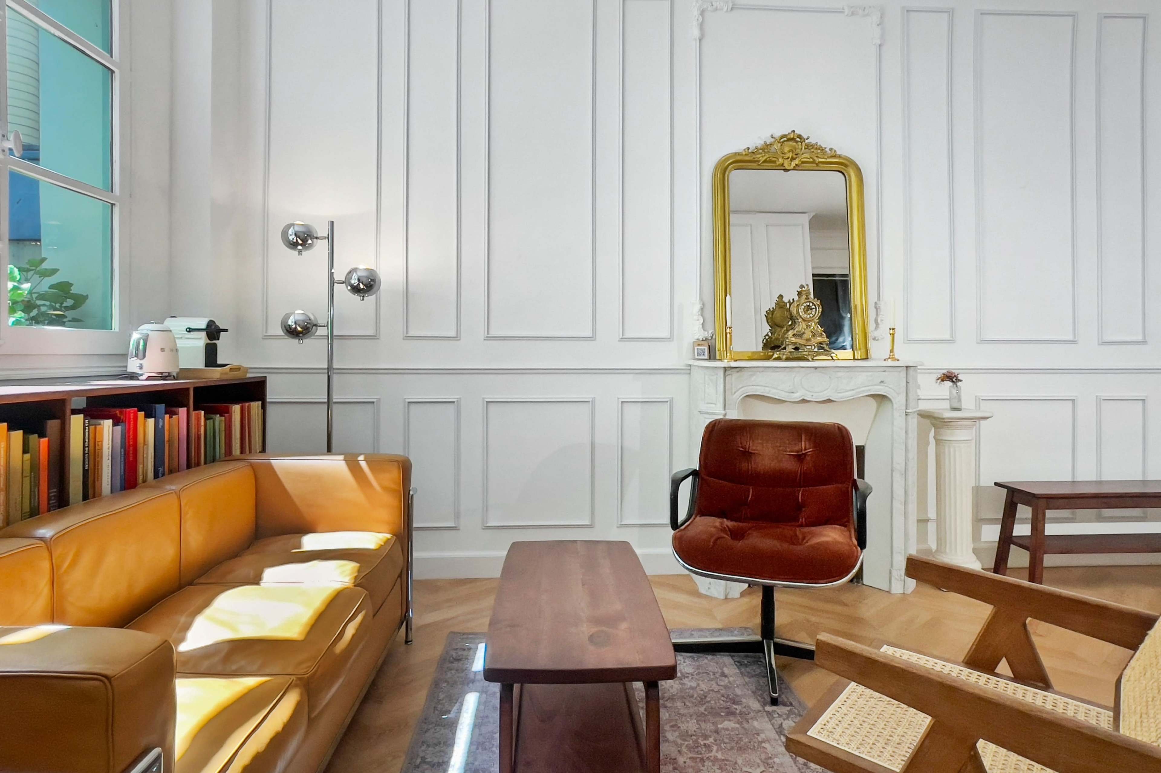 The image shows a bright living room featuring a leather sofa, a brown armchair, a wooden coffee table, and a mirror with a decorative gold frame against white paneled walls.