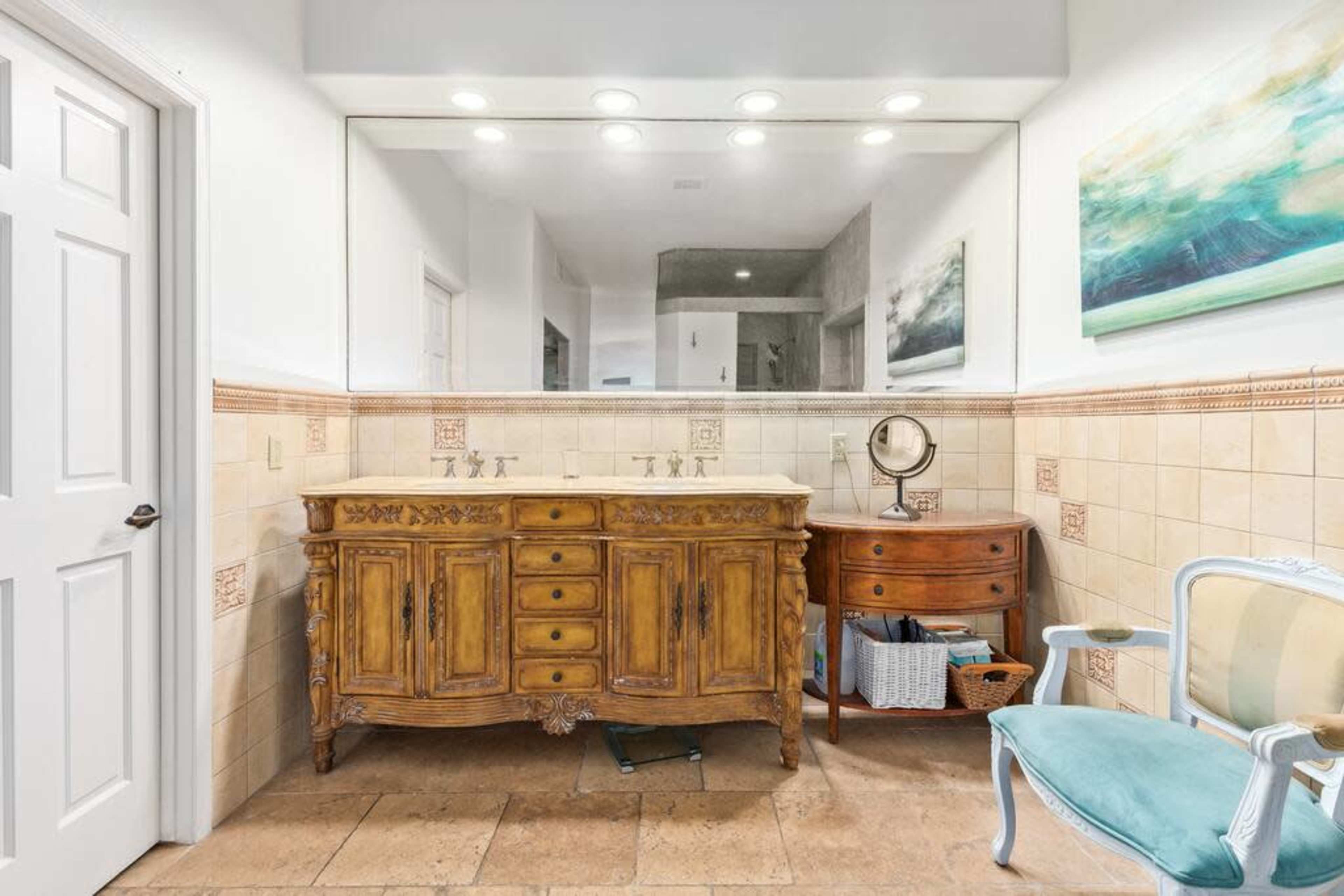 A bathroom featuring a wooden double sink vanity with a large mirror above, surrounded by beige tiled walls and a blue upholstered chair in the corner.