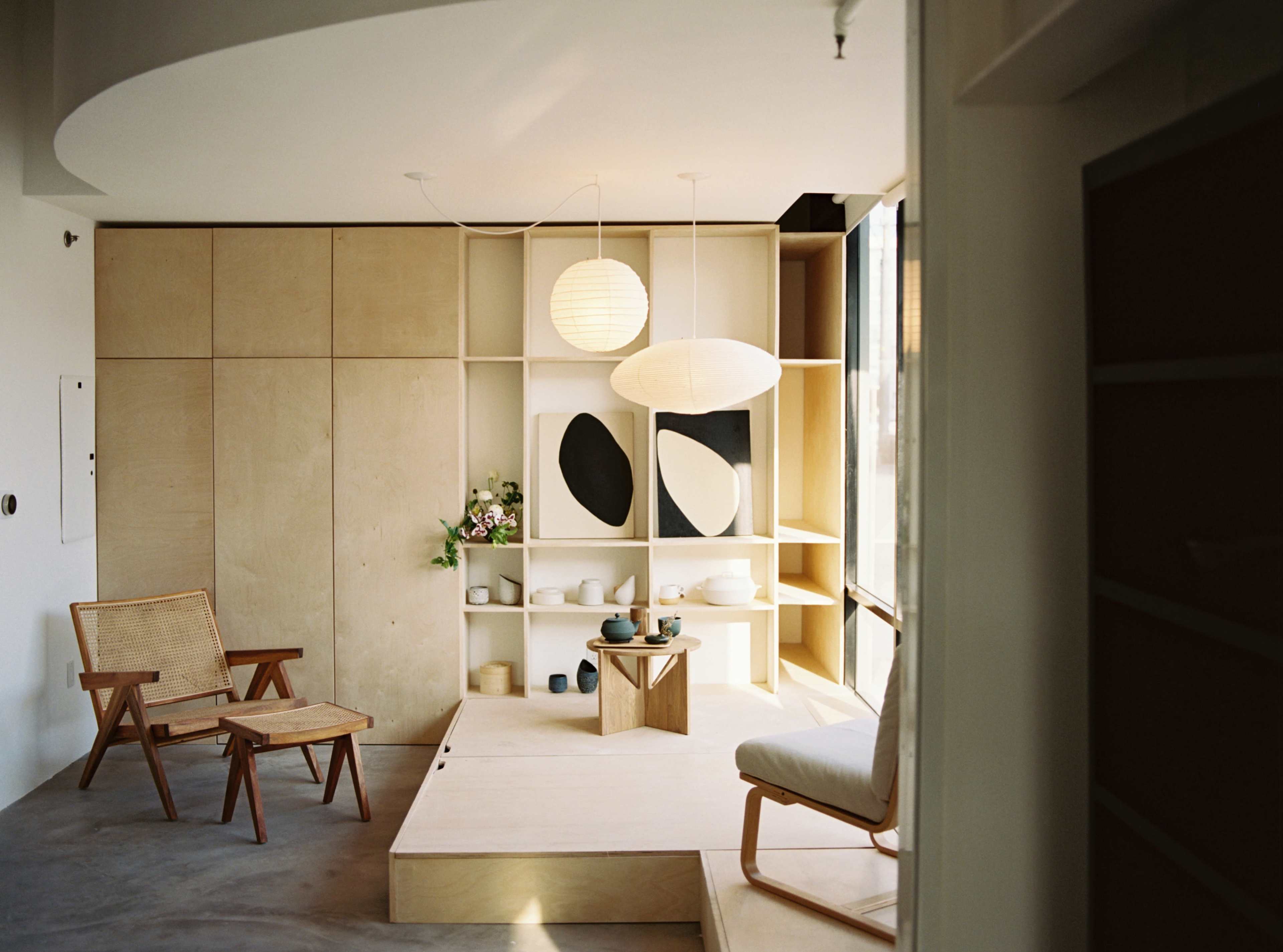 A minimalist interior space featuring a wooden platform, a lounge chair, and a light fixture above a display of artistic decor and pottery.