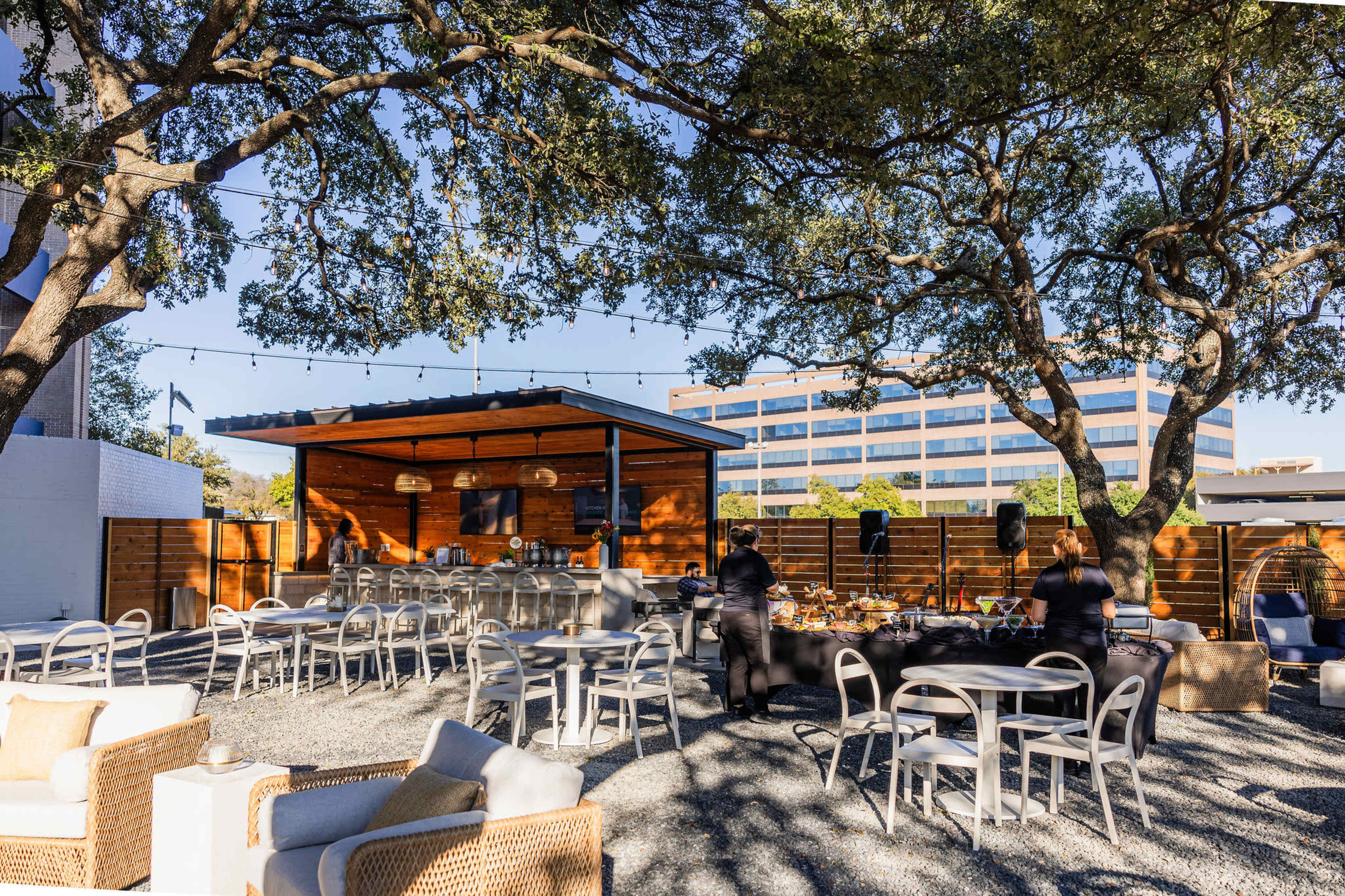 The outdoor space features a wooden structure surrounded by trees, with tables and chairs arranged on a gravel surface.