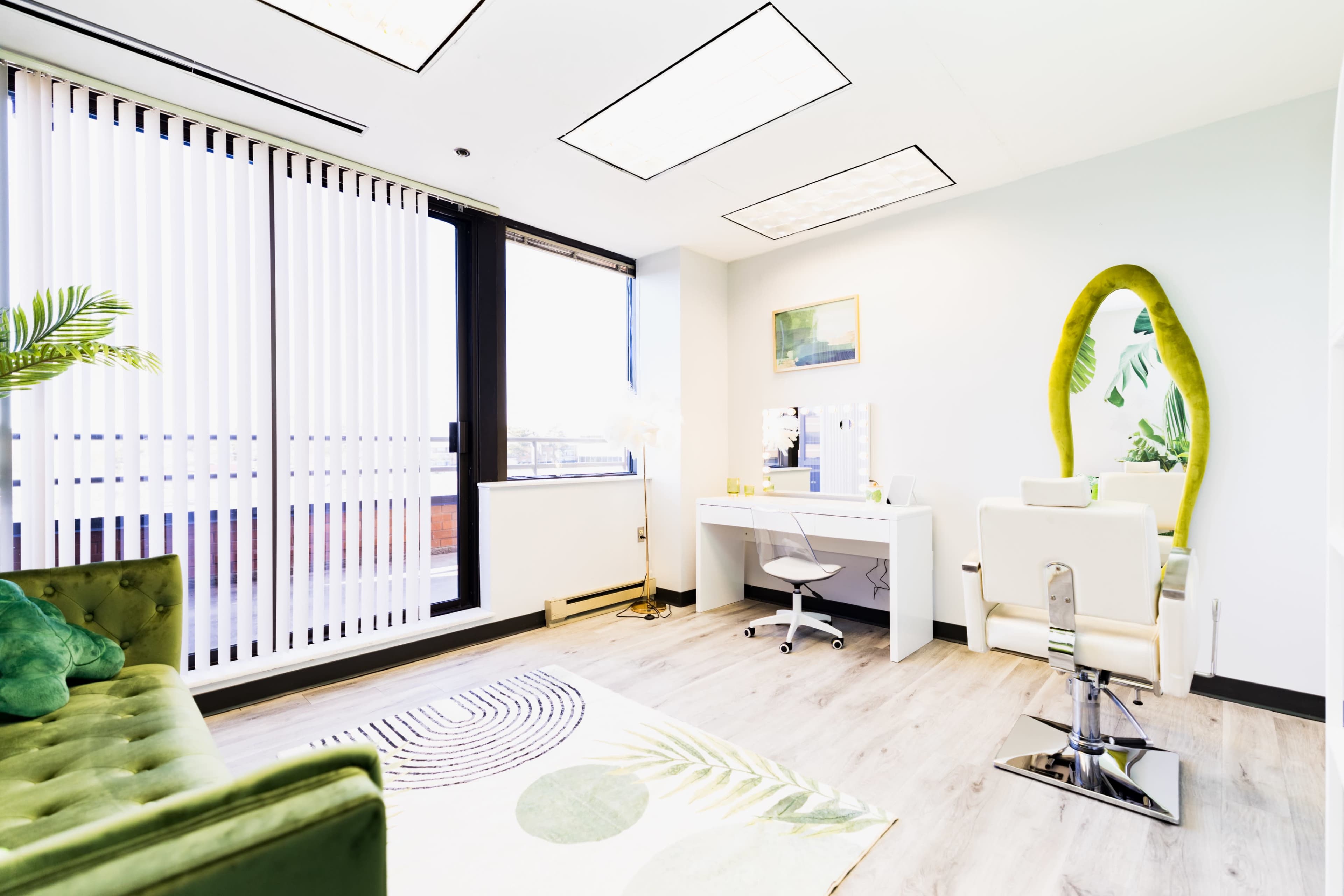 The image shows a modern salon interior with a chair and mirror, a stylist's station, a green couch, and large windows featuring vertical blinds.
