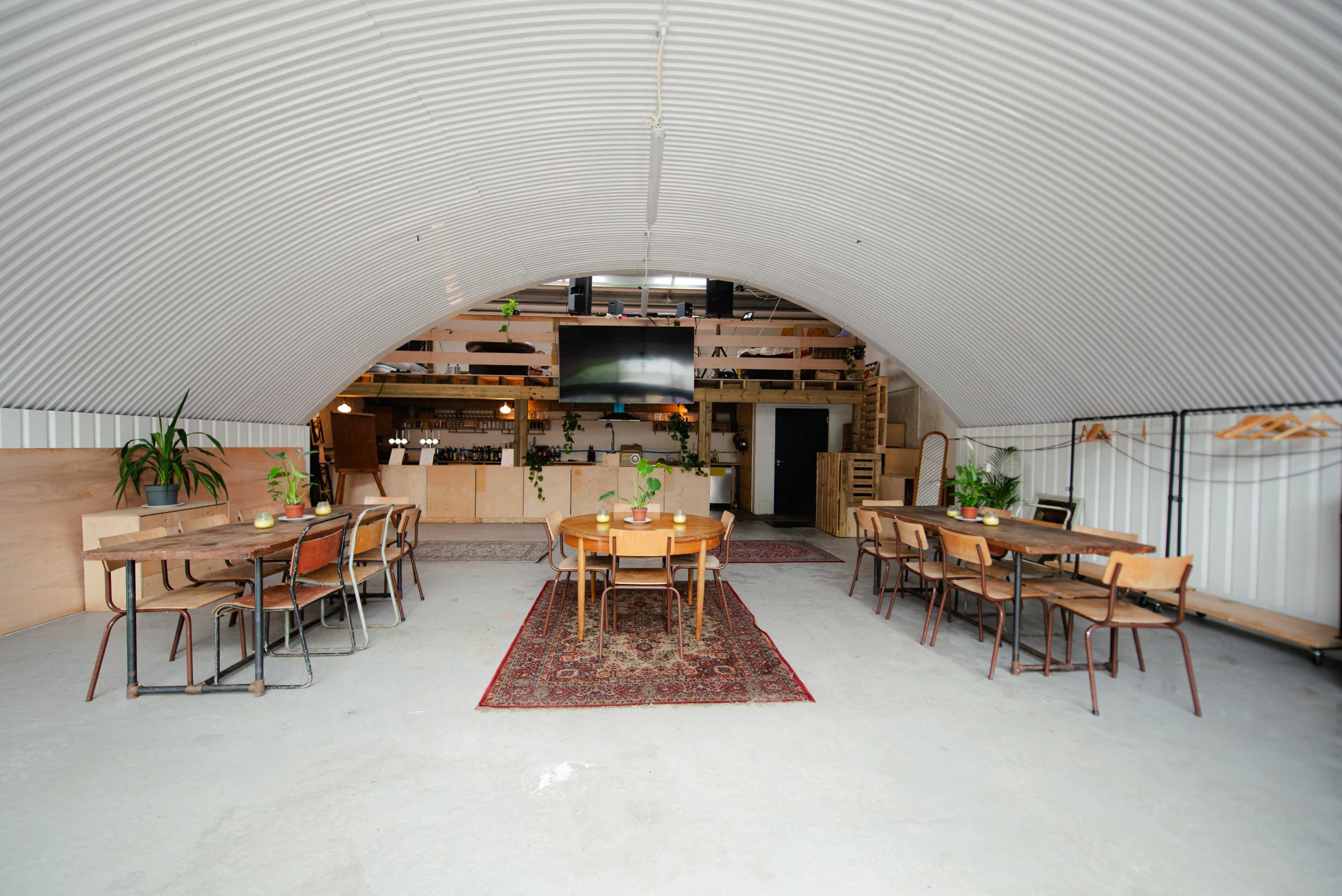 The interior of an industrial-style space features wooden tables and chairs arranged around a central area, with a bar and kitchen setup visible in the background.