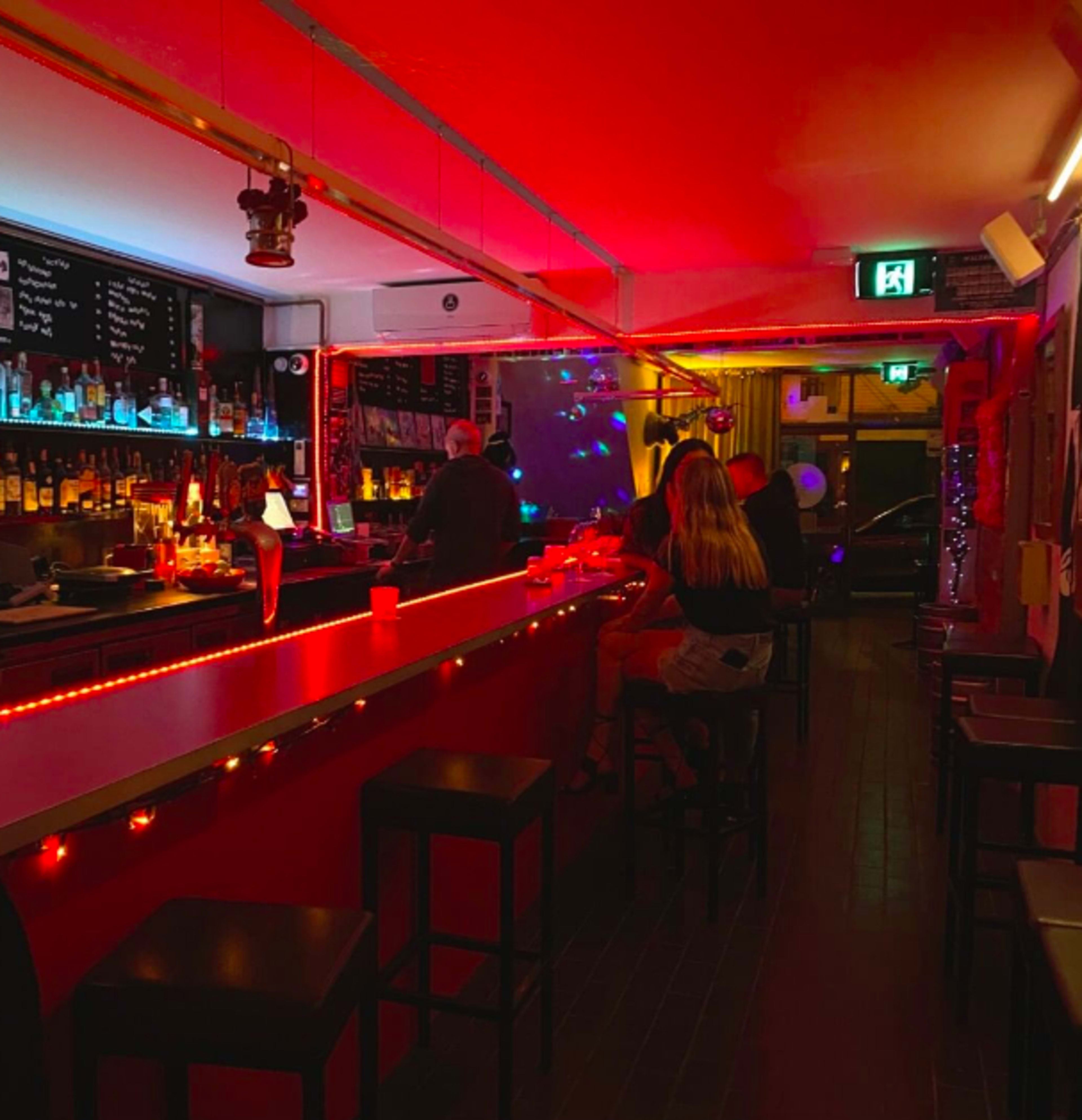 A dimly lit bar with red and blue lighting, featuring a long bar counter and patrons sitting on stools.