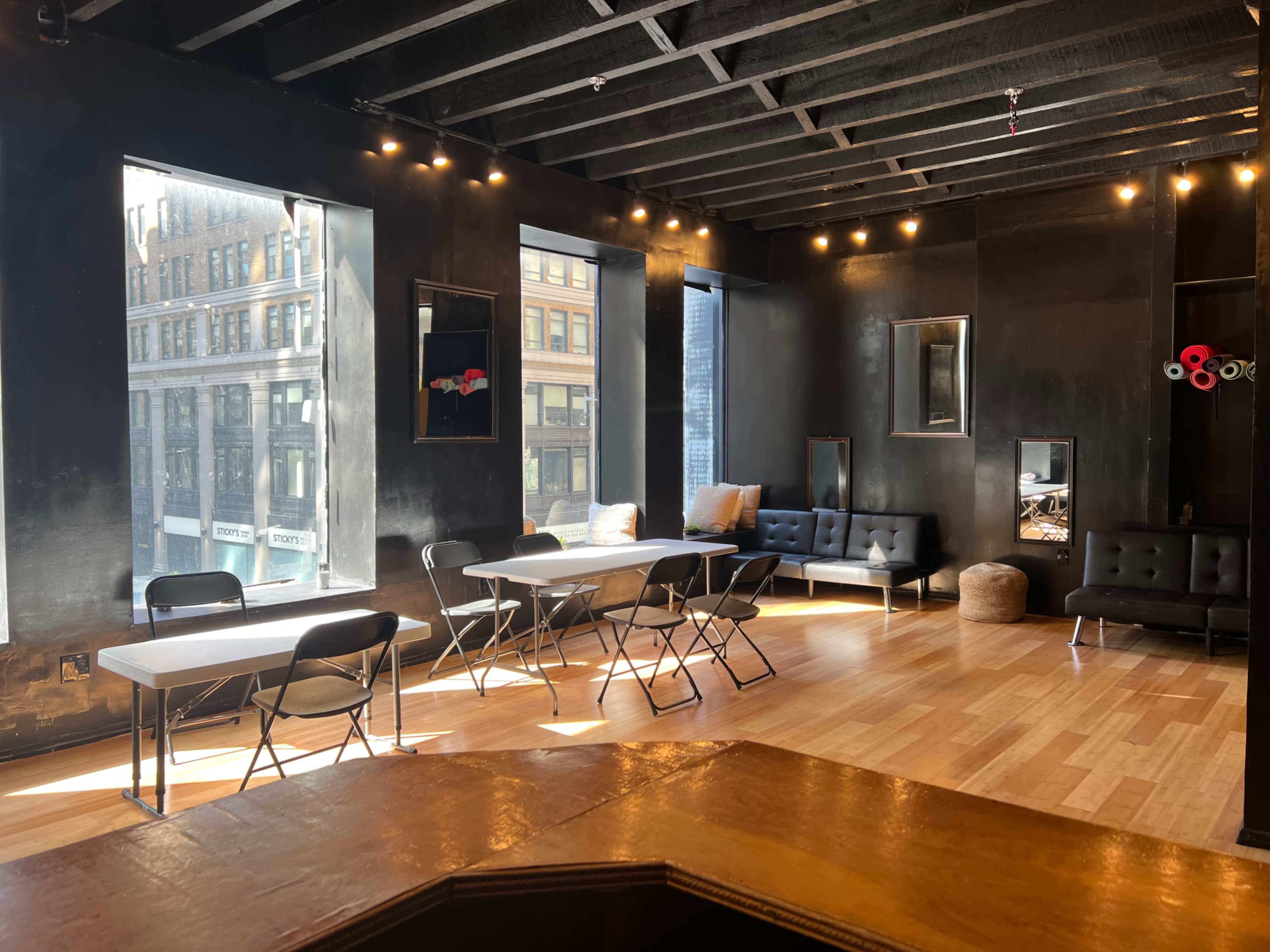 The image shows a modern interior space with large windows, black walls, and folding tables surrounded by seating areas.