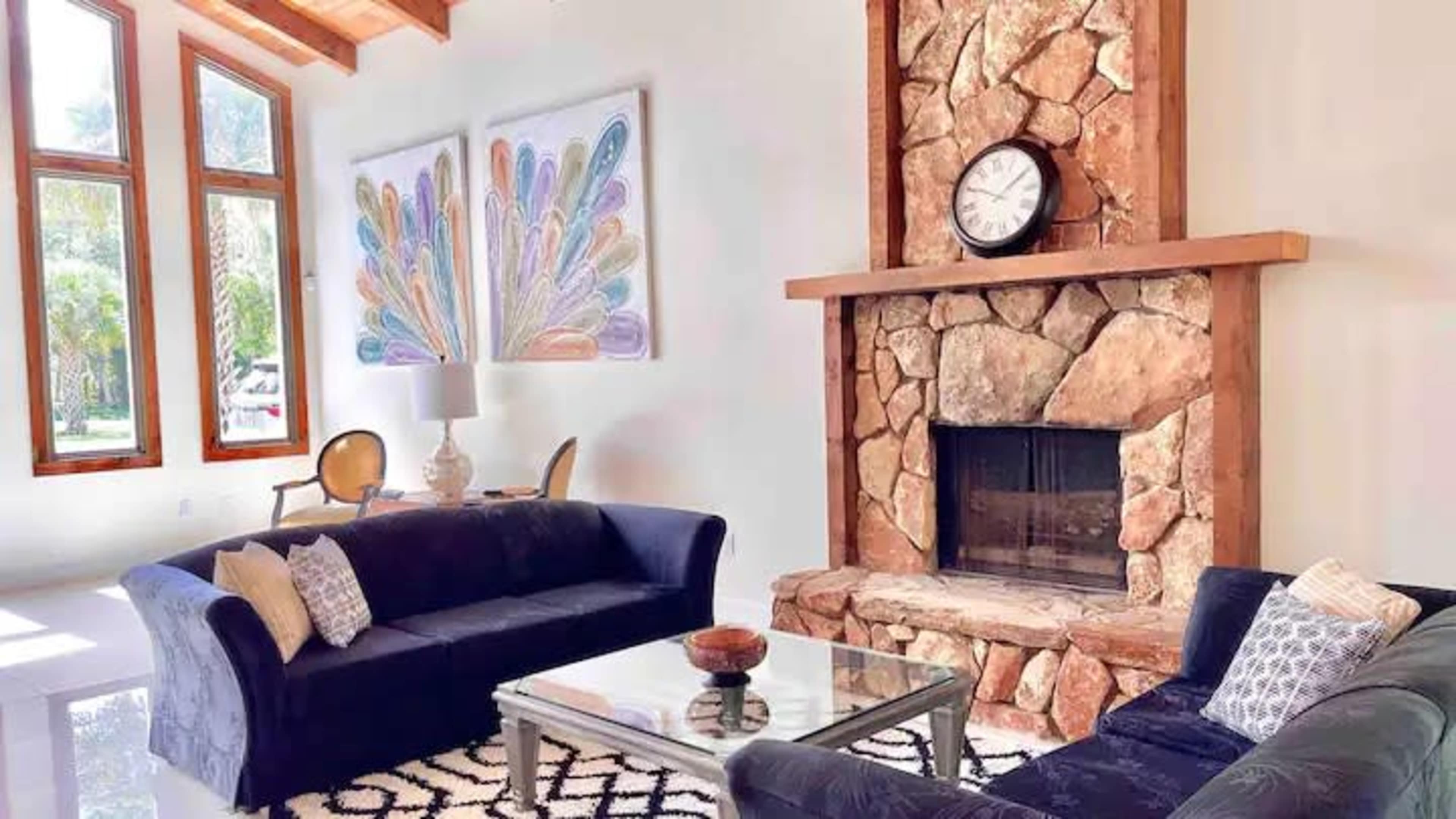 A living room with two dark sofas facing a glass coffee table in front of a stone fireplace, flanked by large windows and colorful artwork on the walls.