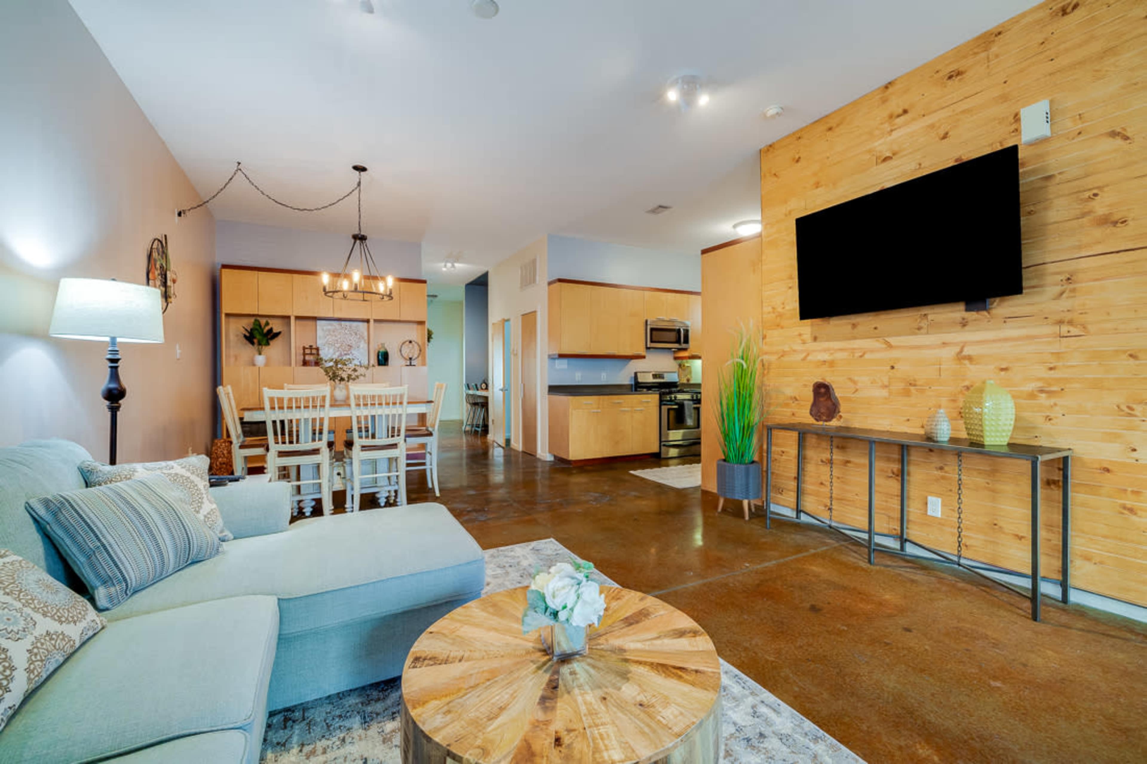 A modern open-concept living area featuring a light blue sofa, a wooden coffee table, a dining area with a large table, and a kitchen in the background, all complemented by a wooden accent wall.
