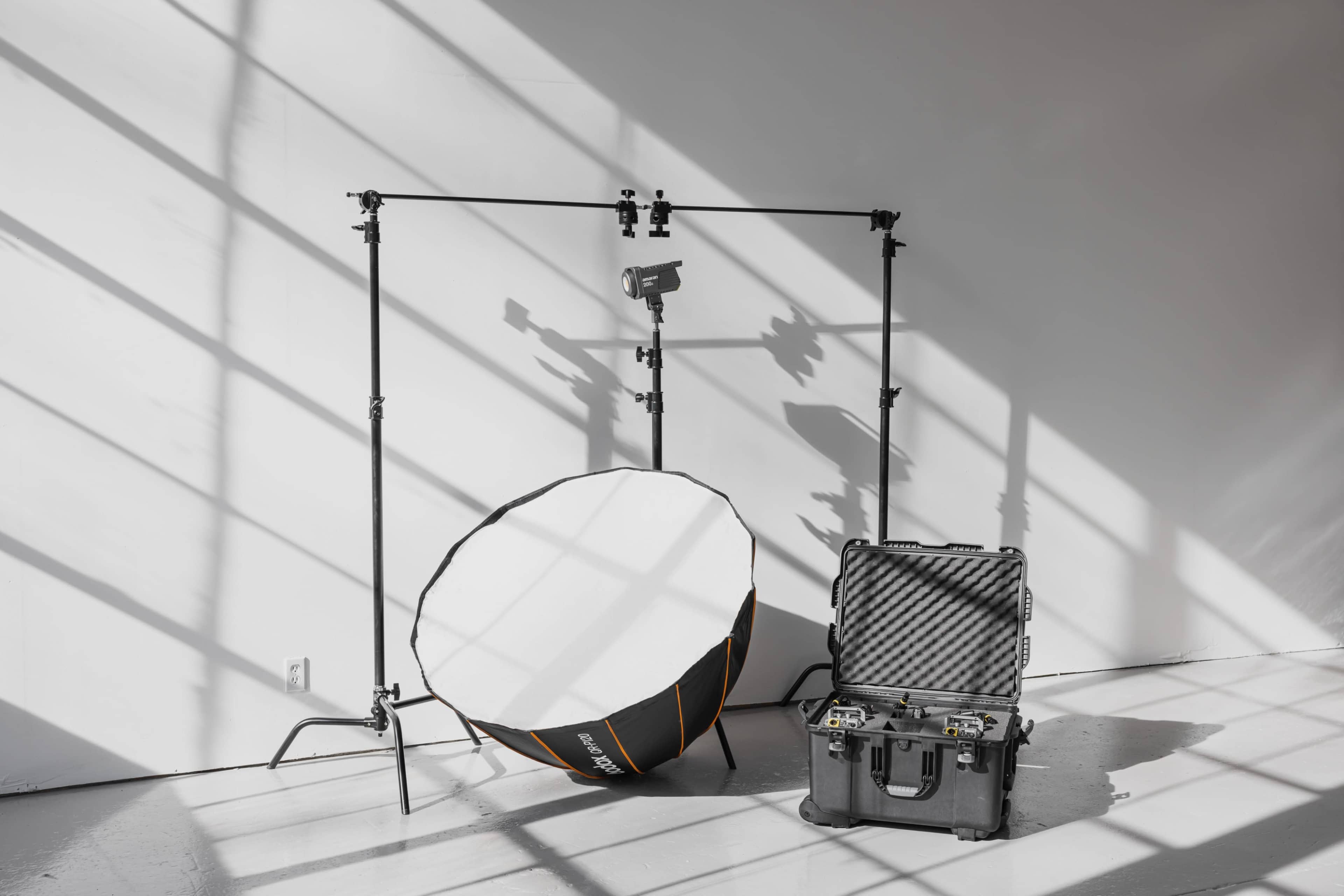 The image shows a photography setup featuring a large softbox on a stand, a light mounted above, and a sturdy black case on the floor, all placed in a studio with sunlight casting shadows on the wall.