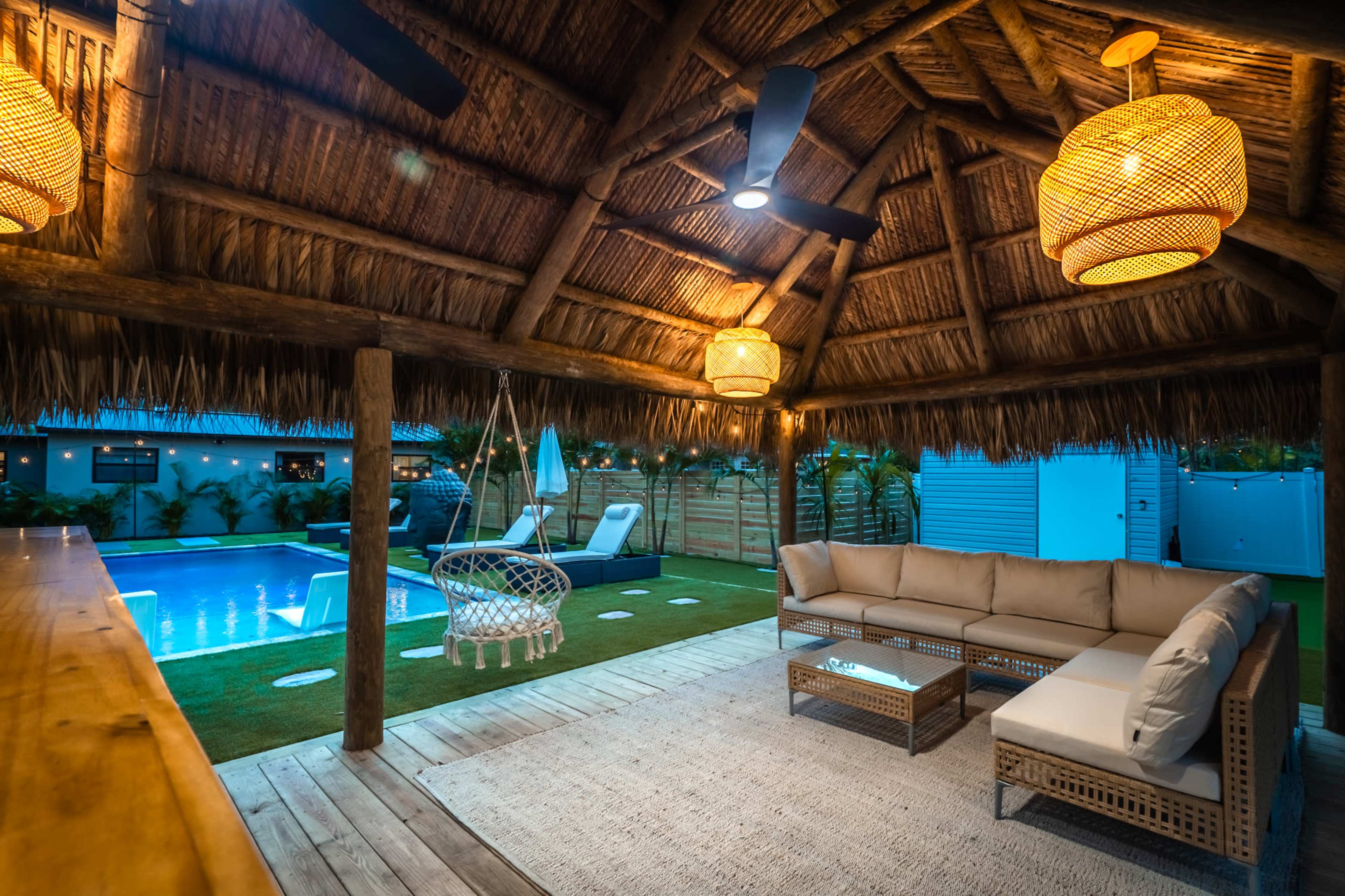 A thatched-roof outdoor lounge area features a sectional sofa, hanging chair, and pool with lighting in the background.
