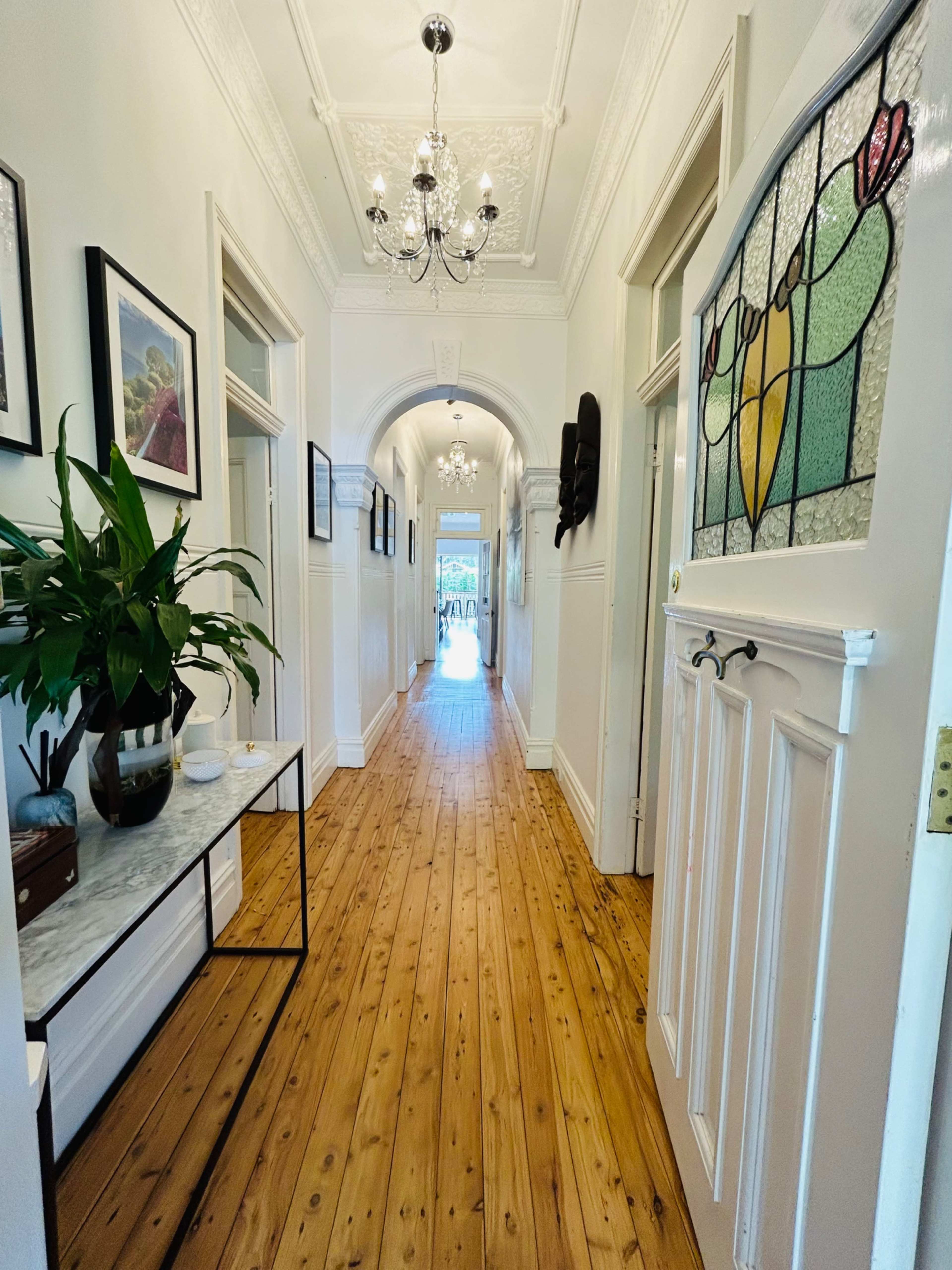 A long hallway with wooden flooring, framed artwork on the walls, a potted plant, and a door featuring stained glass.