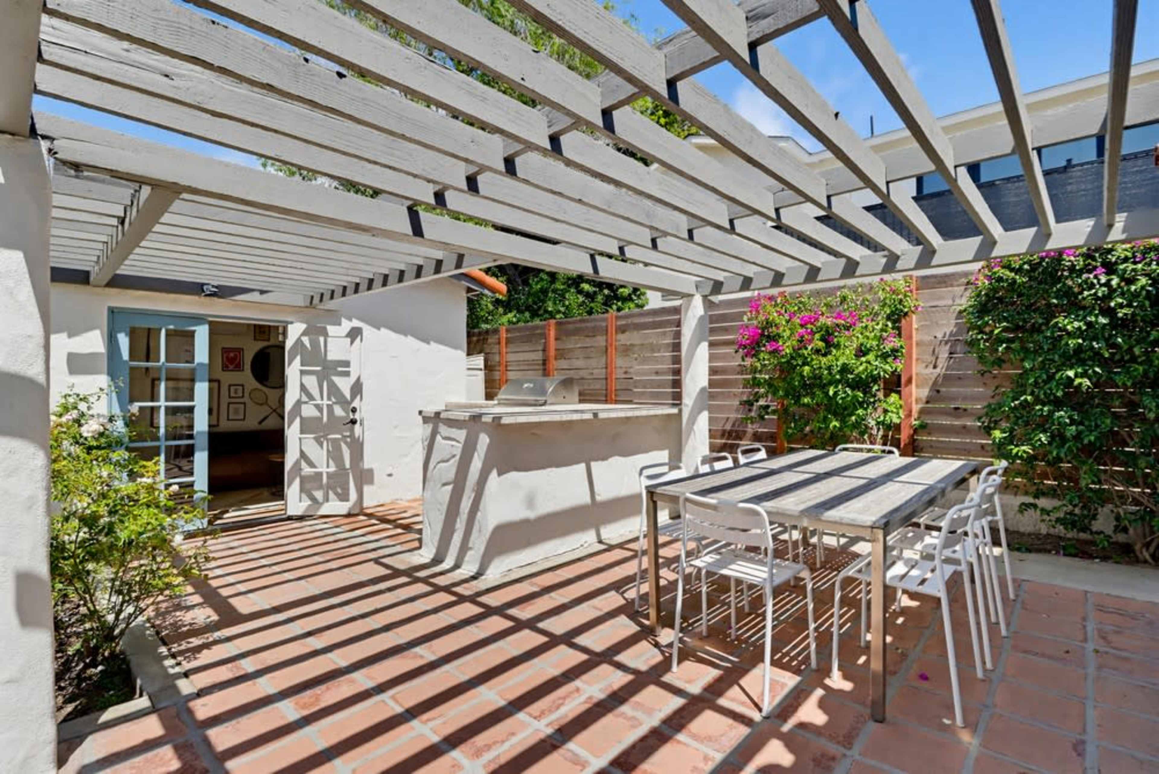 The image shows an outdoor patio area with a wooden pergola, a bar counter, and a dining table surrounded by greenery.