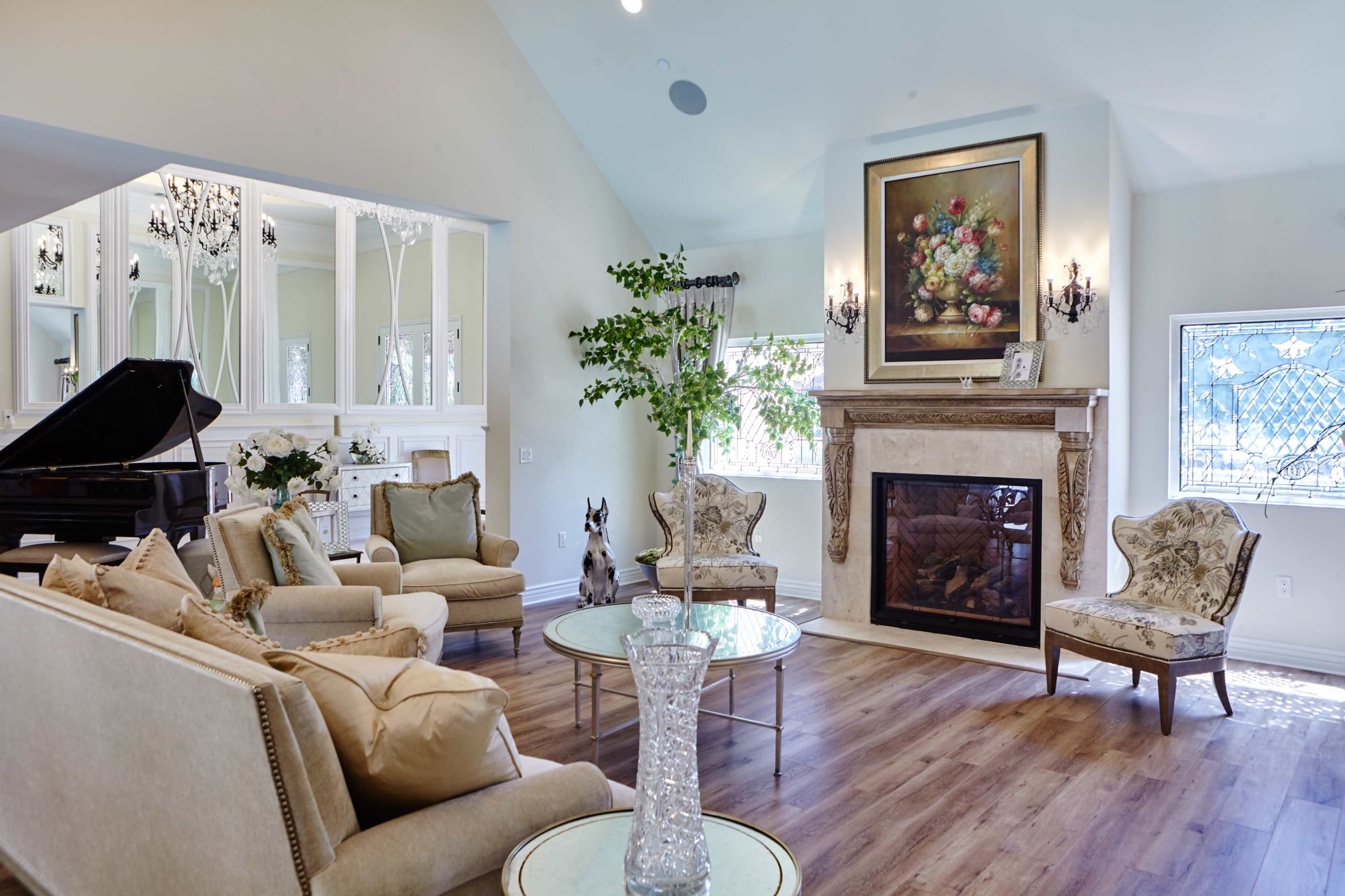 The image shows a bright, elegantly furnished living room with a piano, a fireplace, and comfortable seating arranged around a glass coffee table.