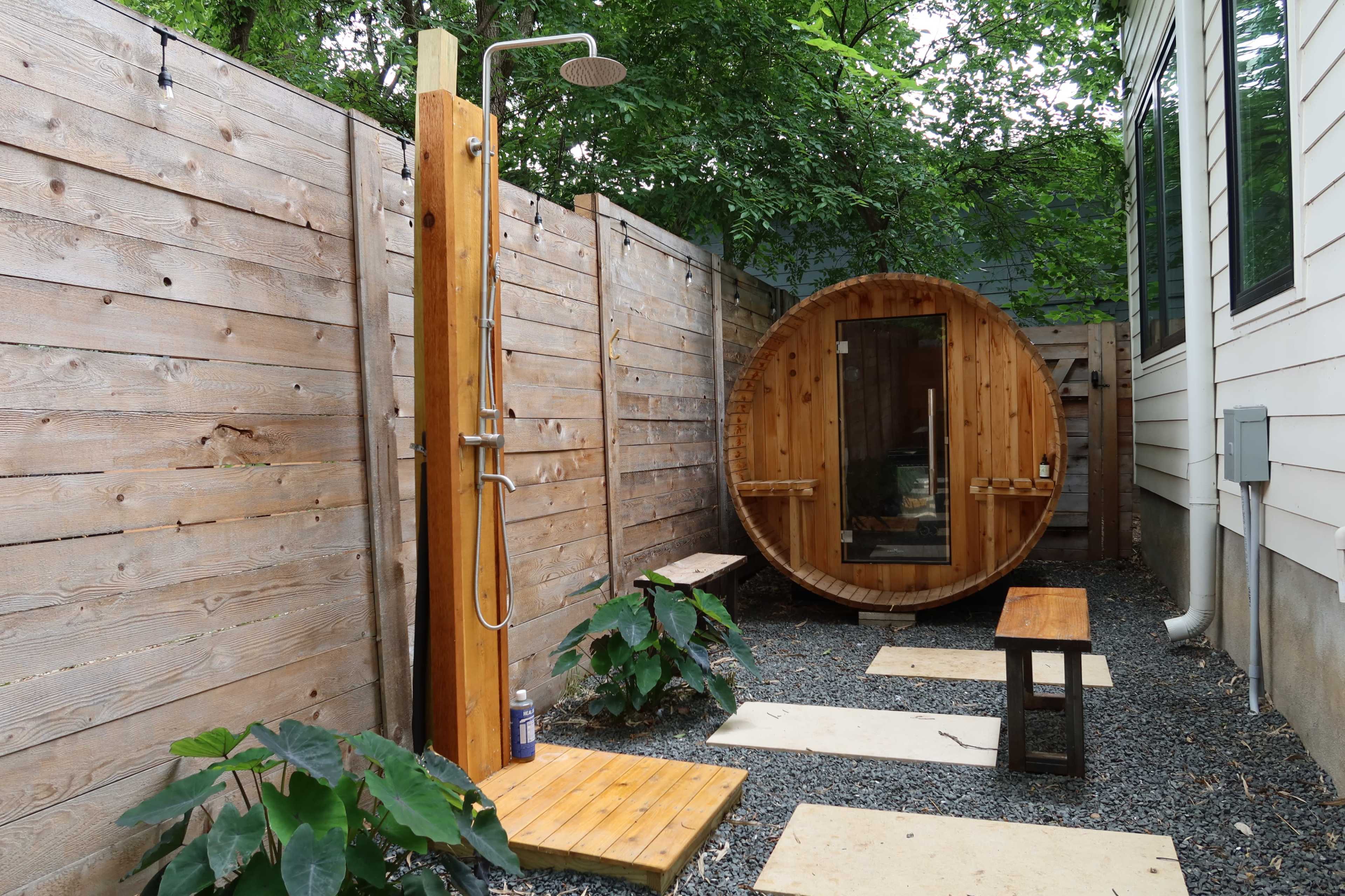 A wooden shower next to a round sauna, surrounded by a gravel path and bordered by wooden fences and plants.