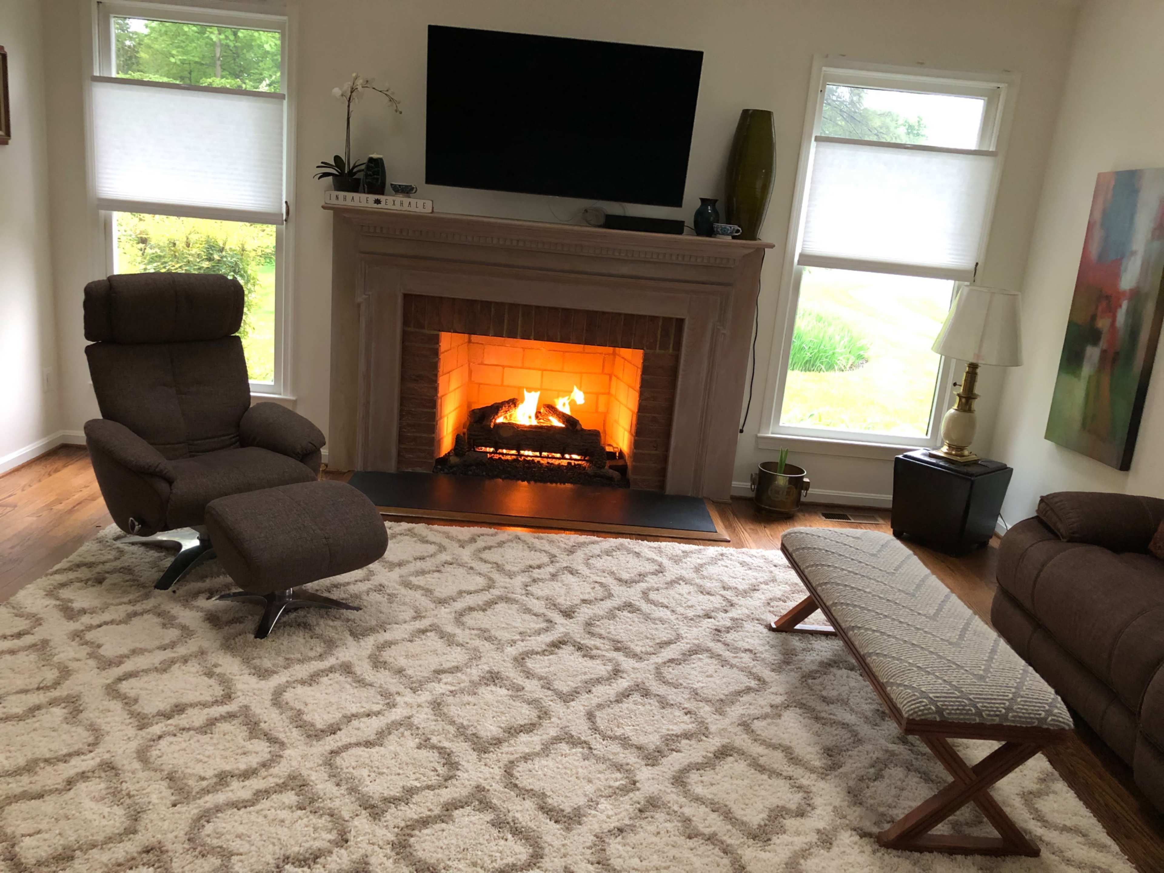 The image shows a cozy living room with a fireplace, two chairs, a bench, a large rug, and a television mounted on the wall.