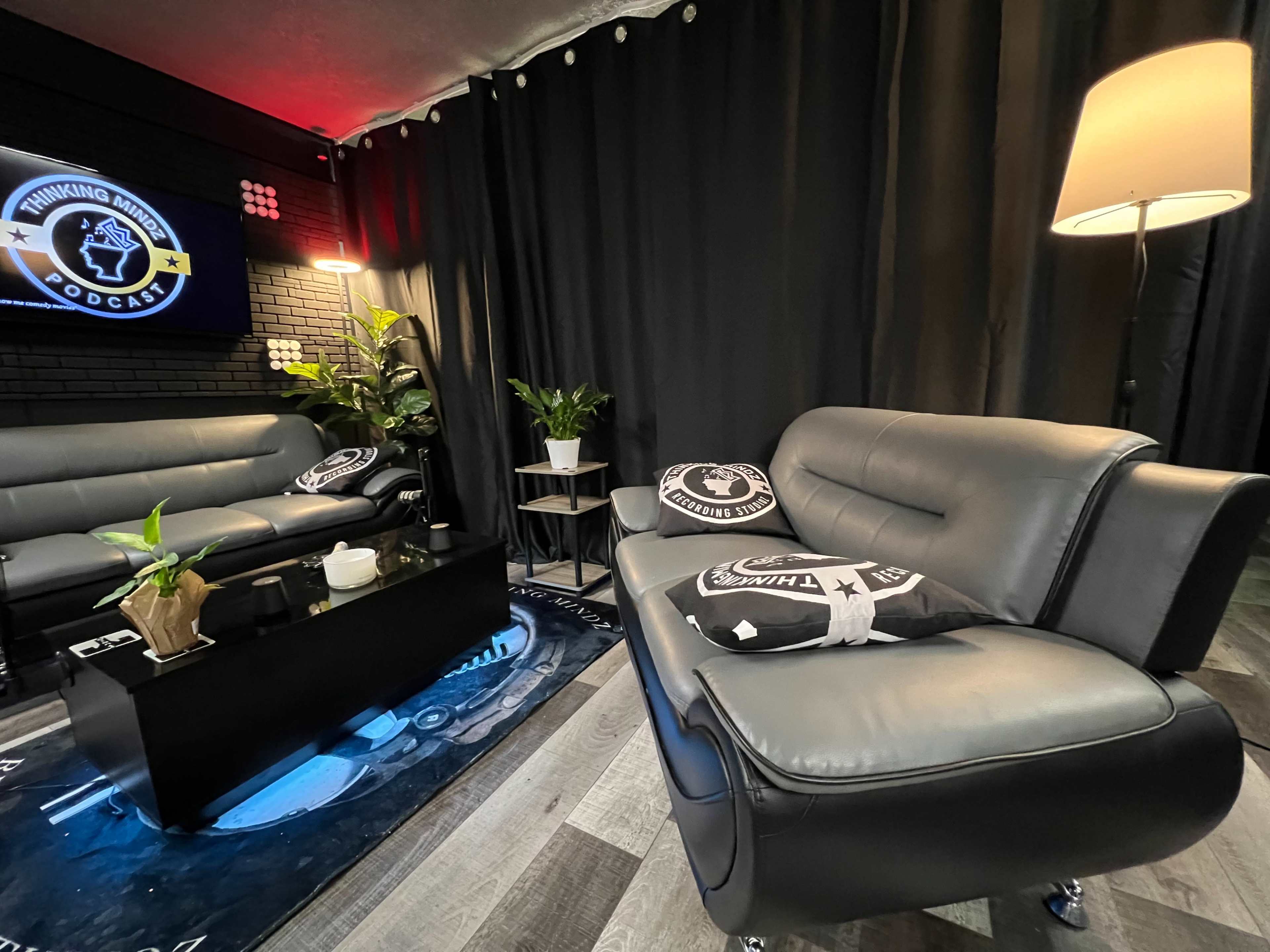 A modern lounge area with a black backdrop, a coffee table, two leather sofas, a floor lamp, and potted plants, designed for a podcast studio.