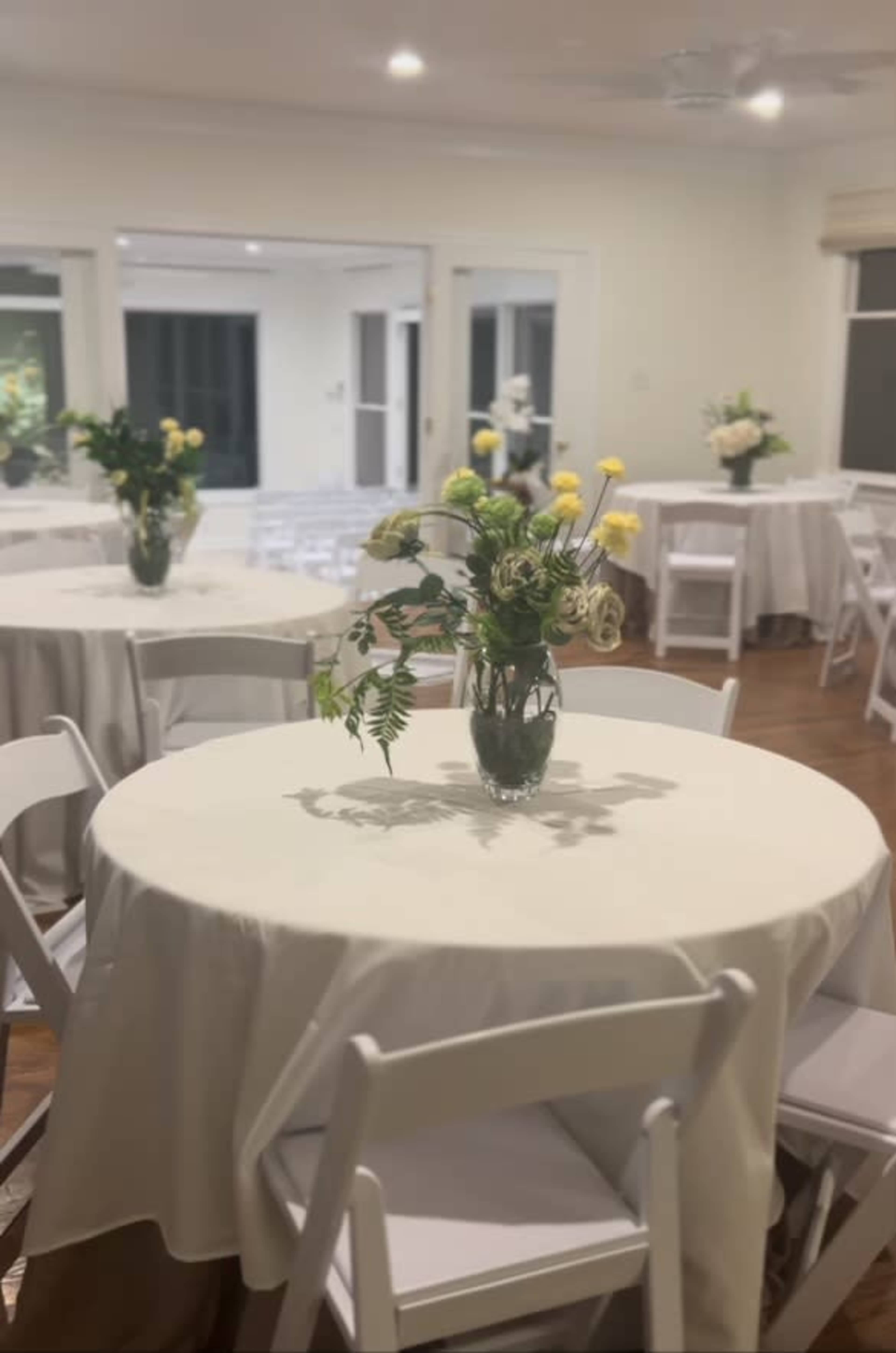 The scene shows a brightly lit event space with round tables covered in white tablecloths, each featuring a vase of flowers at the center.