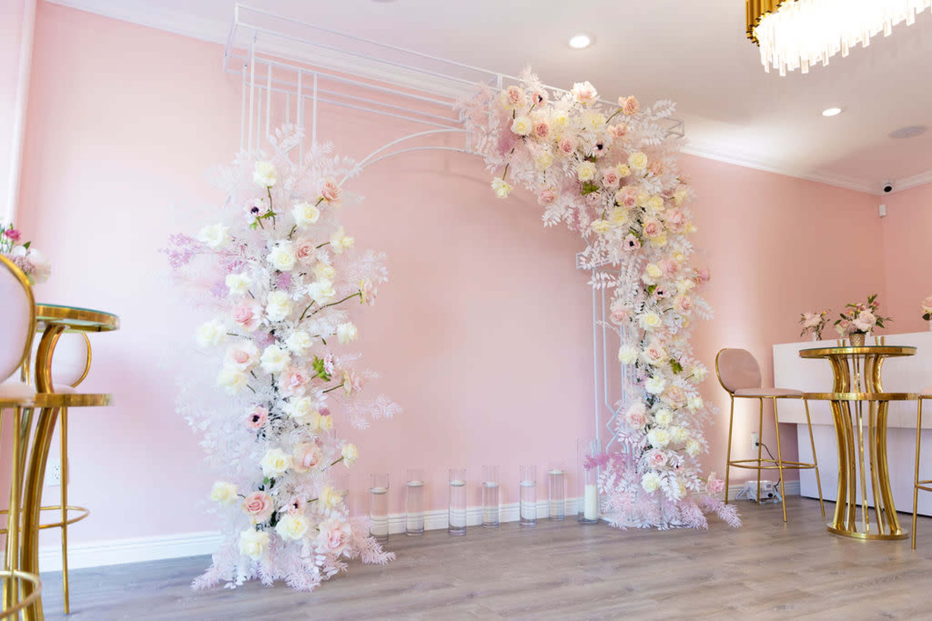 The image shows a floral arch decorated with pastel flowers and greenery against a pink wall, accompanied by gold furniture and a table.