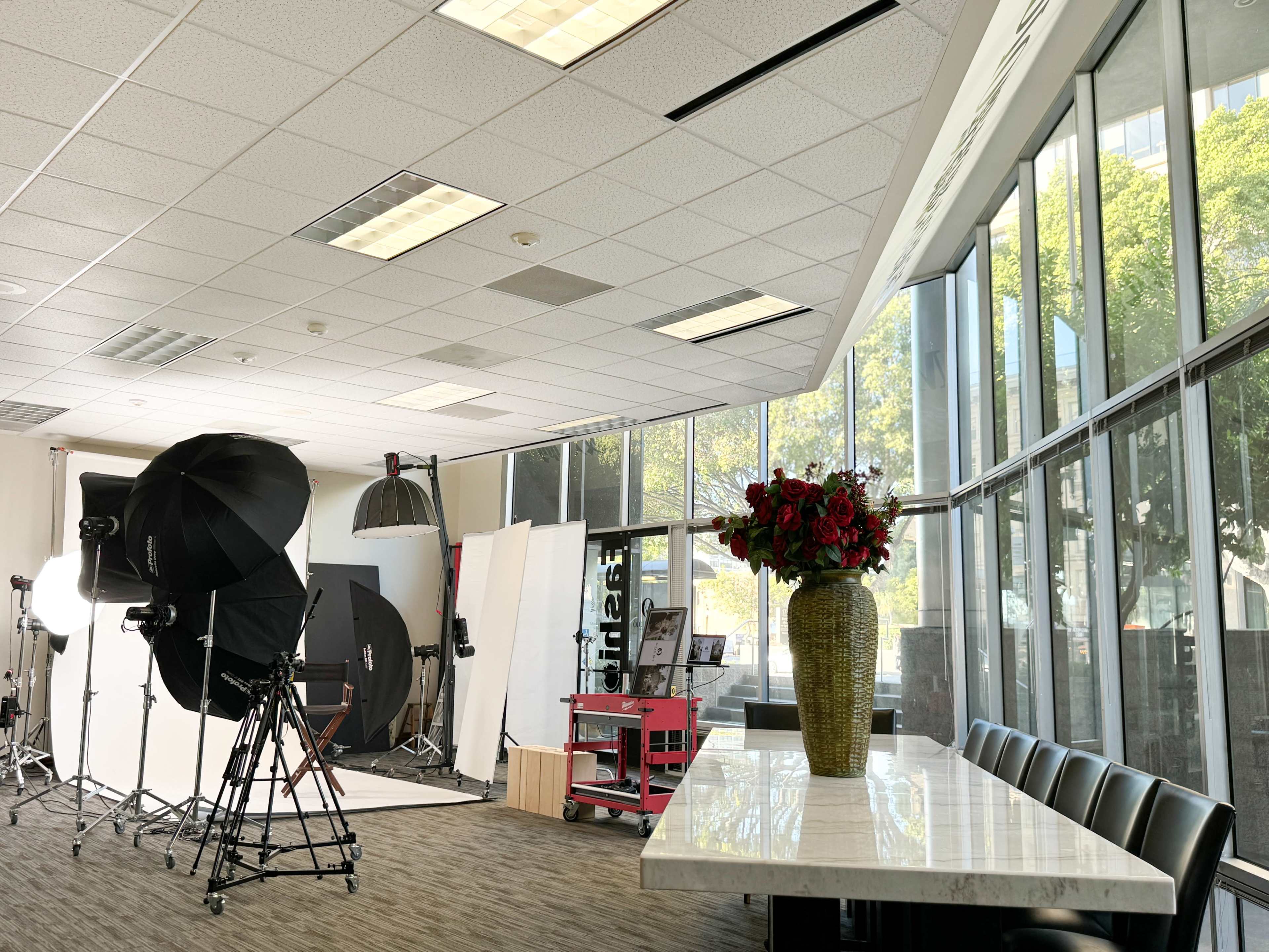 The image shows a photography studio with equipment and a large window, featuring a table with a vase of flowers.