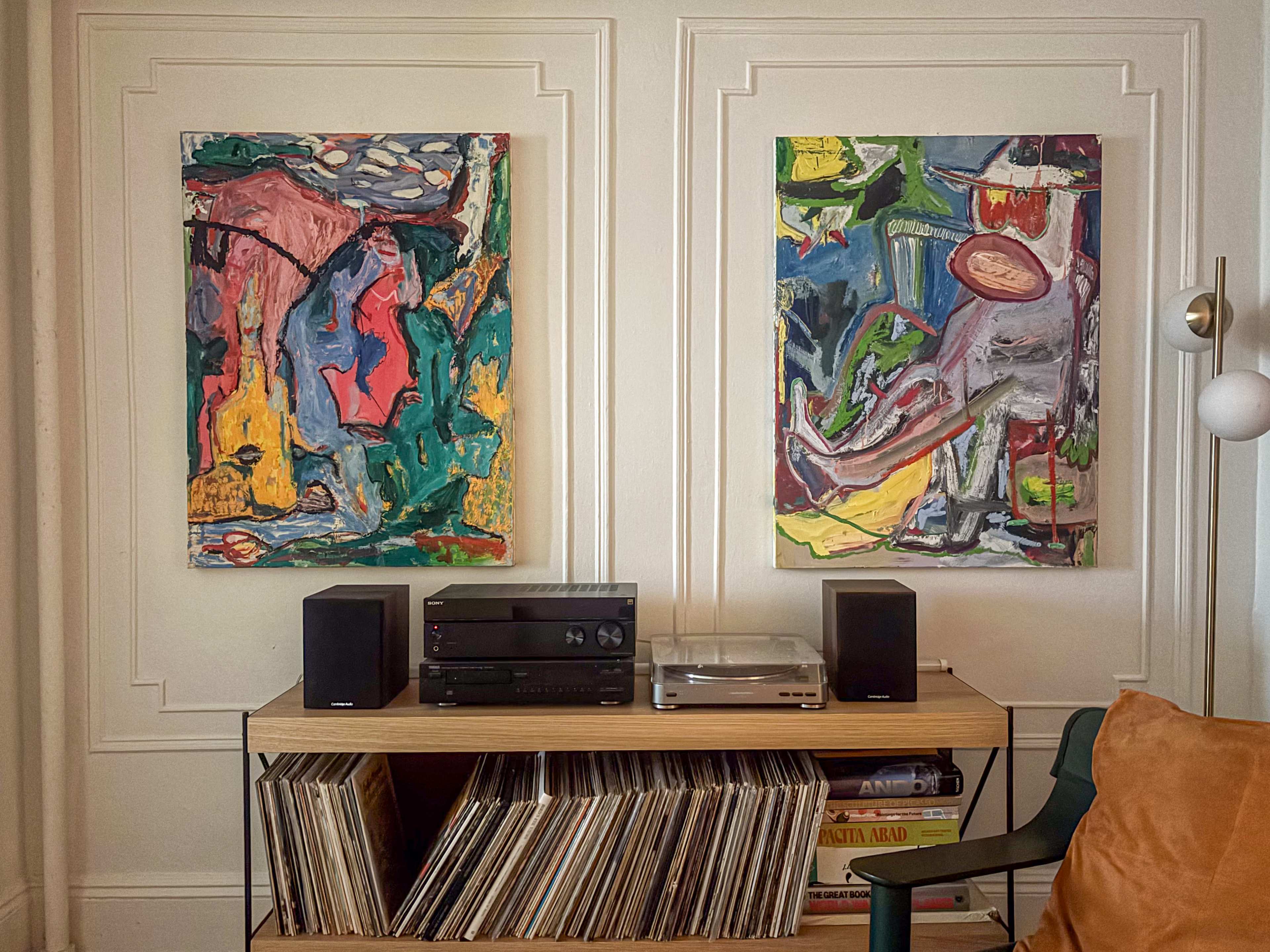 The image shows a wooden shelf with vinyl records, a turntable, and speakers, flanked by two colorful abstract paintings on the wall.