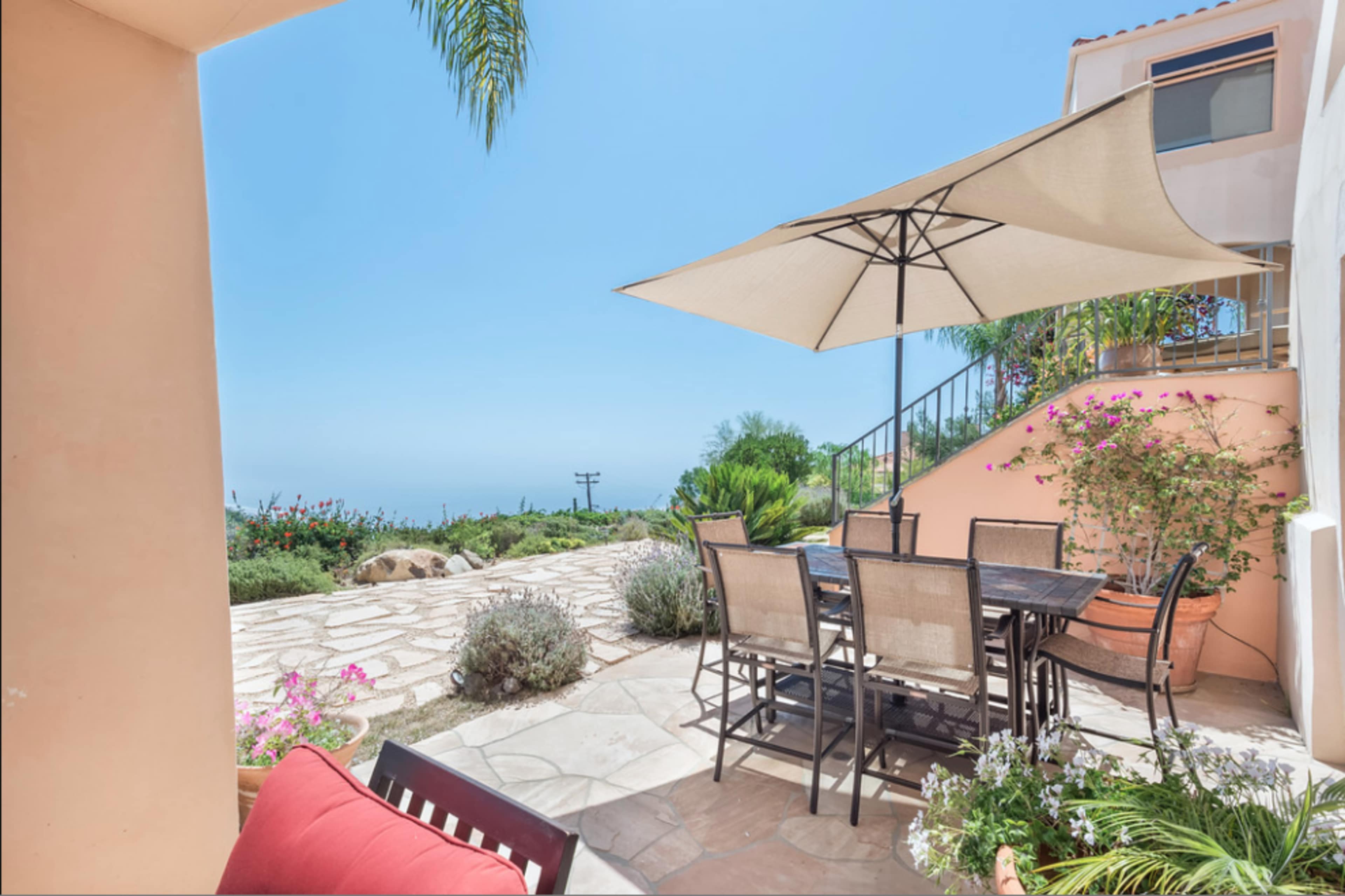 A shaded outdoor dining area with a table and chairs overlooks a stone pathway and lush greenery.