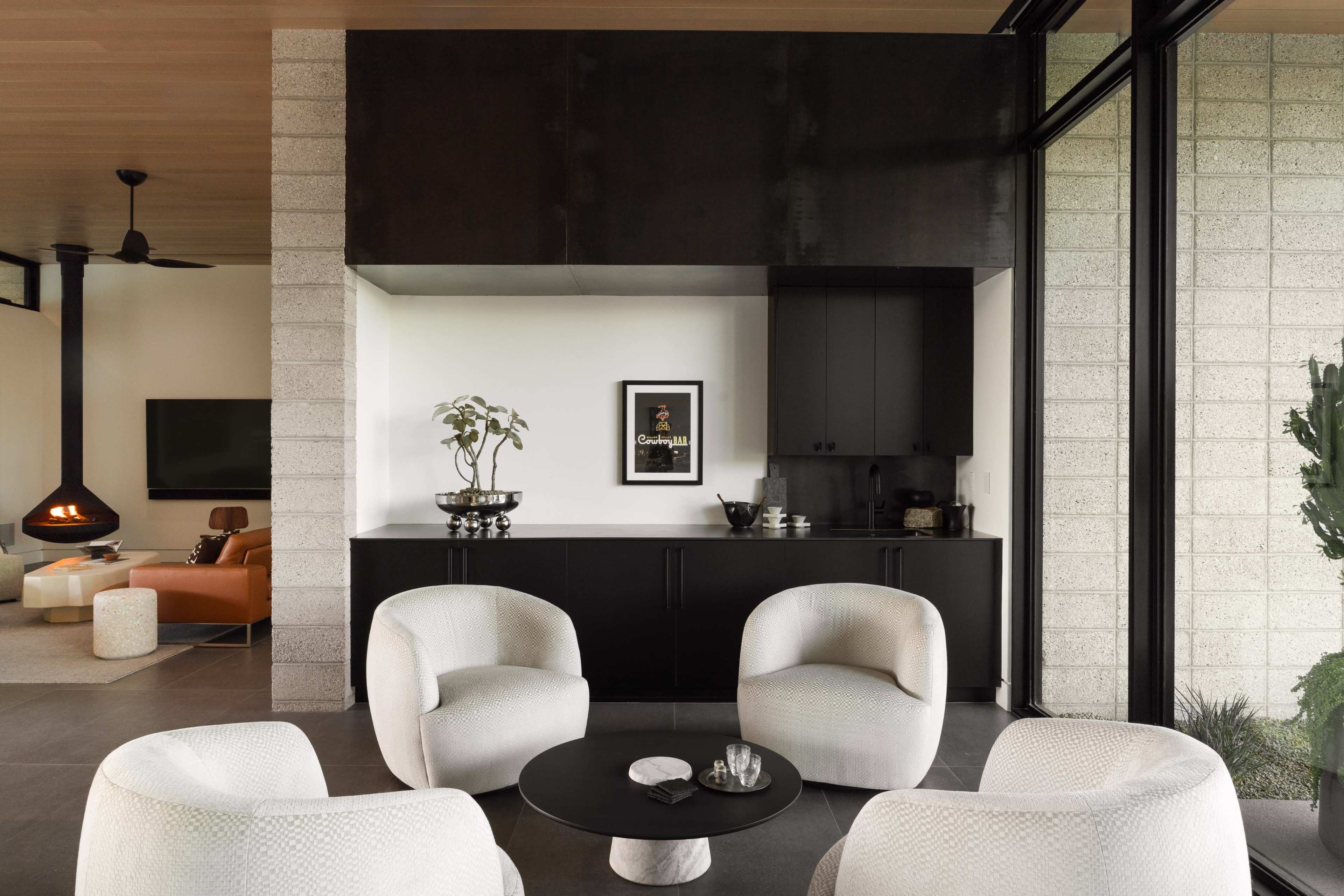 The image shows a modern living space featuring four white chairs arranged around a small black table, with a dark cabinetry wall, a plant, and a large window overlooking greenery.