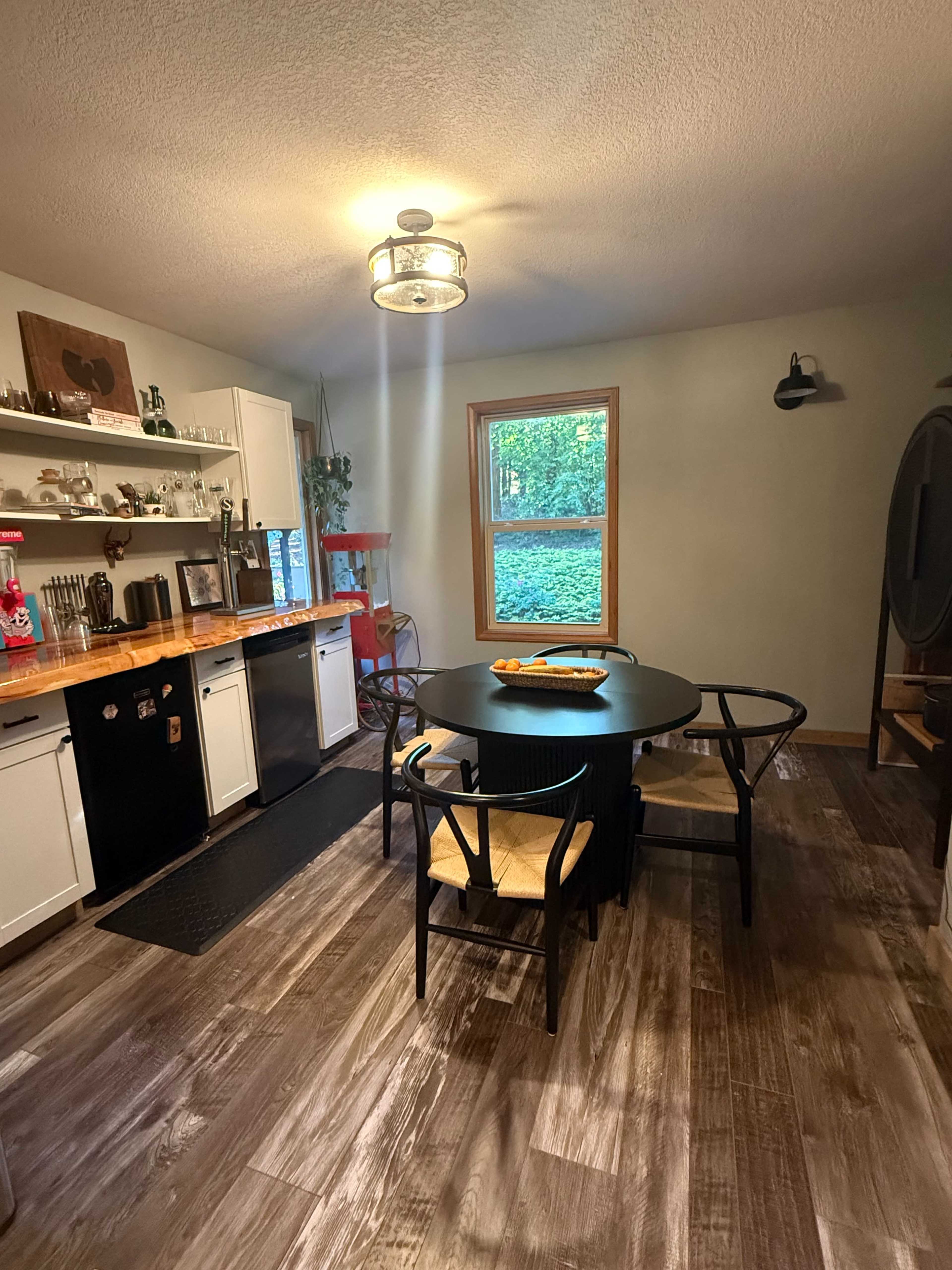 A compact kitchen features a round black dining table with four chairs, hardwood flooring, and large windows overlooking a green outdoor space.