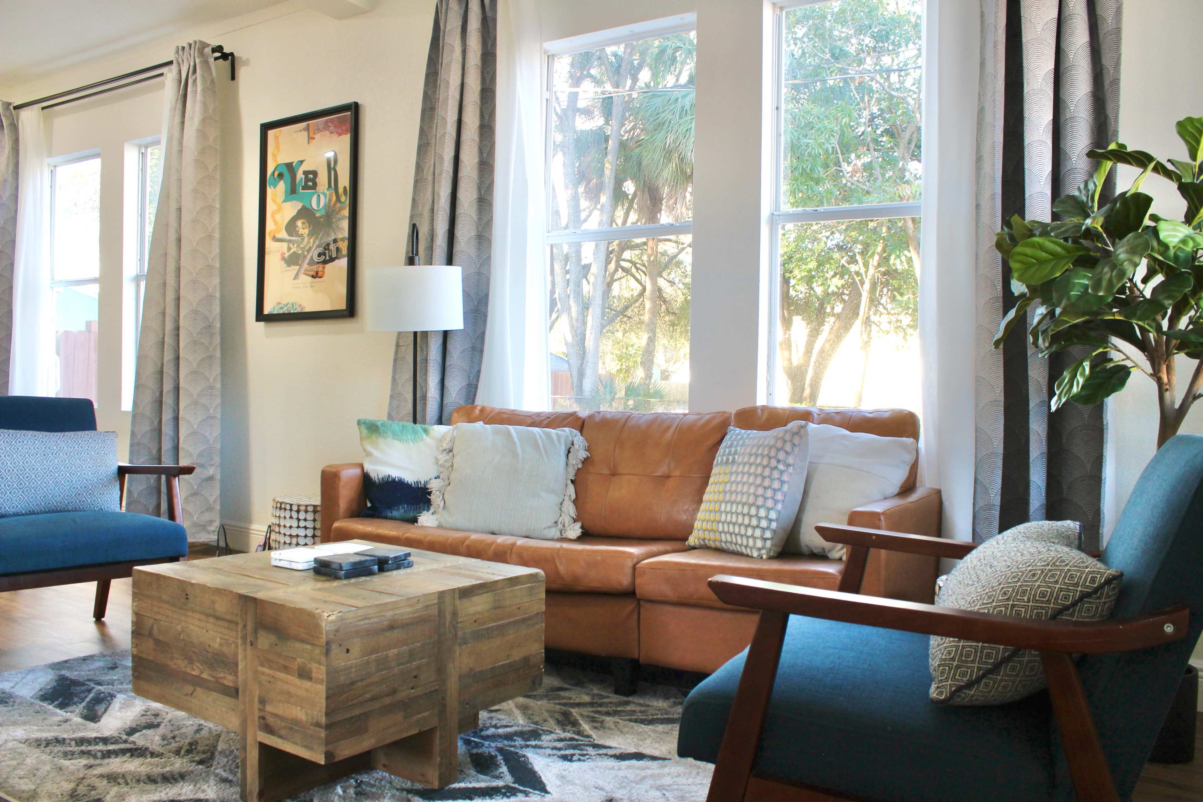 A living room features a brown leather sofa, blue accent chairs, a wooden coffee table, and large windows letting in natural light.