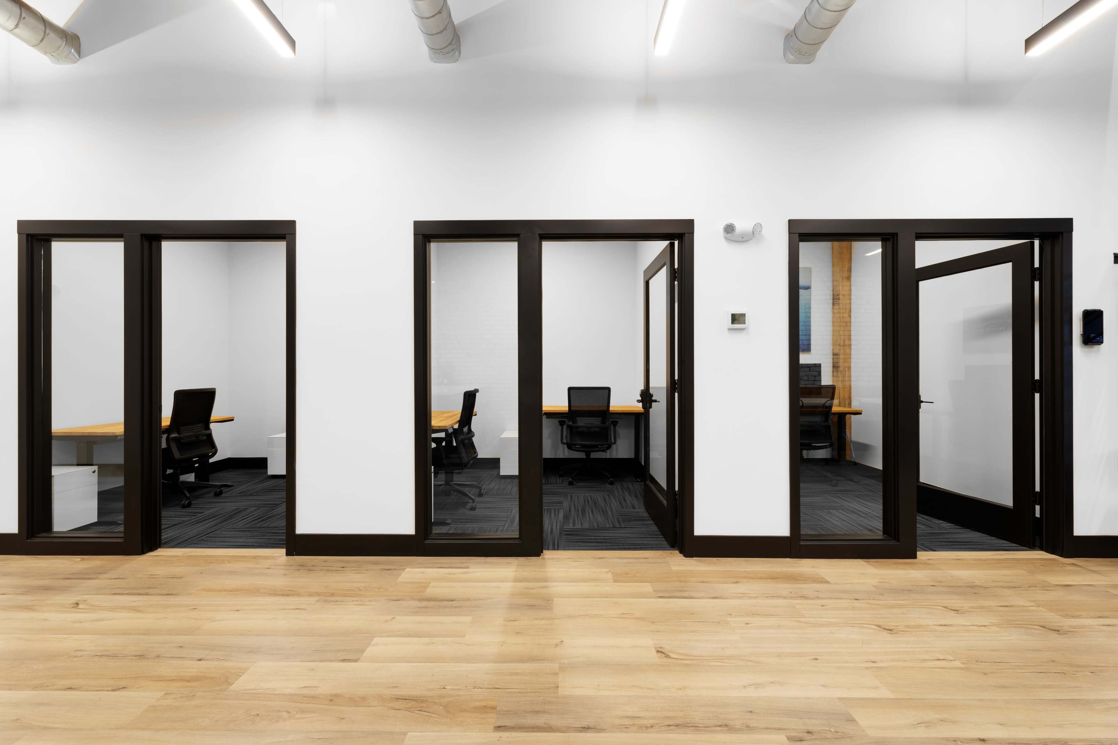 A modern office space with multiple glass-walled meeting rooms and wooden flooring.