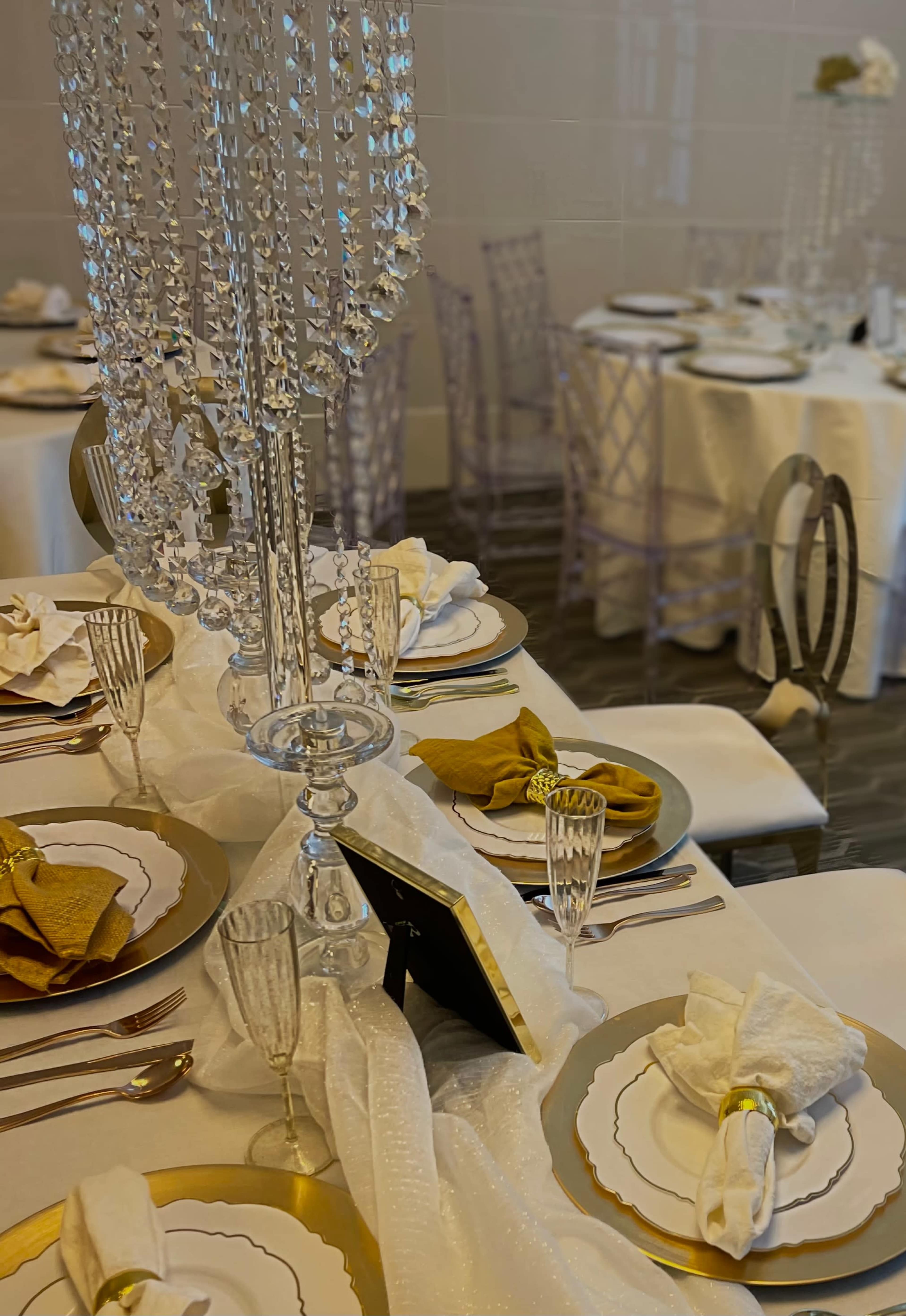 A white table is elegantly set with gold-trimmed plates, crystal glassware, and a decorative chandelier centerpiece.