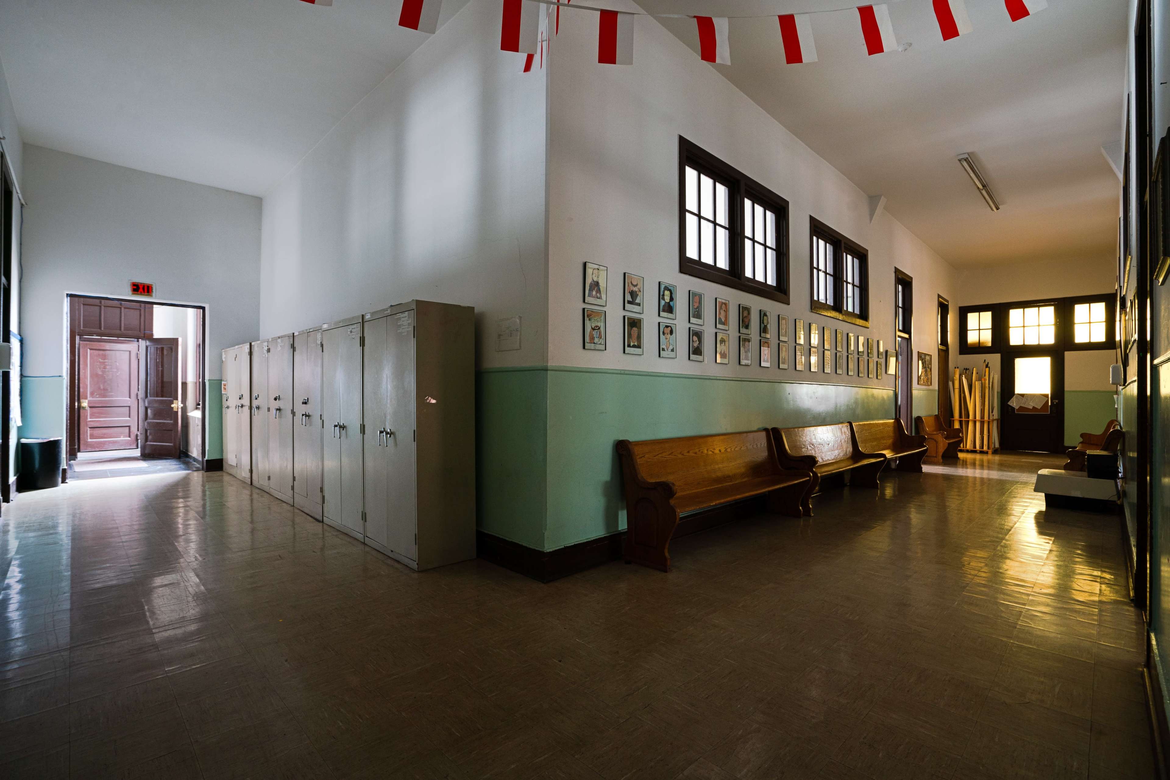 A narrow school hallway features lockers along one wall, benches on the opposite side, and framed photographs hanging on the walls.