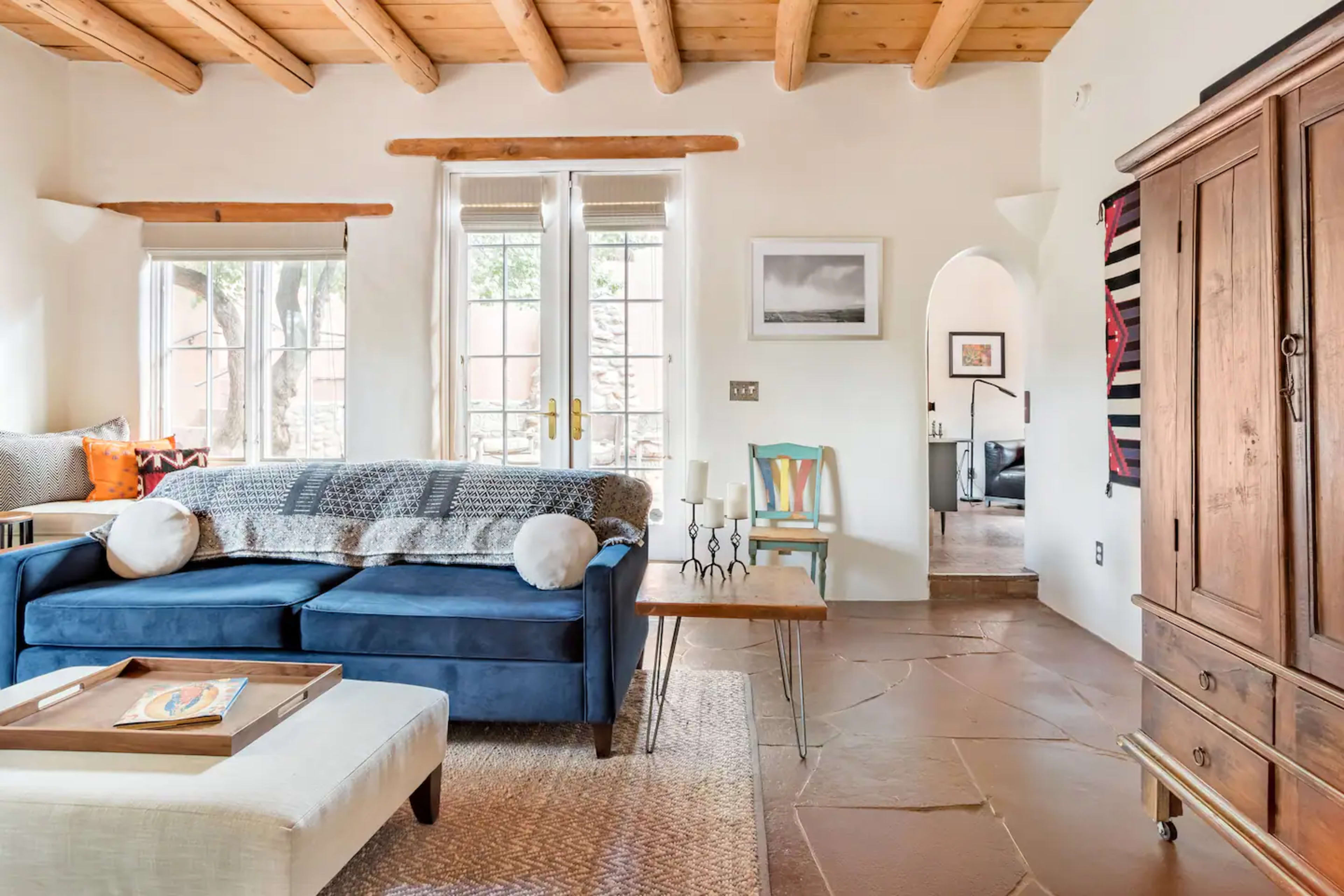 A cozy living room featuring a blue sofa, a wooden table, and large windows allowing natural light.