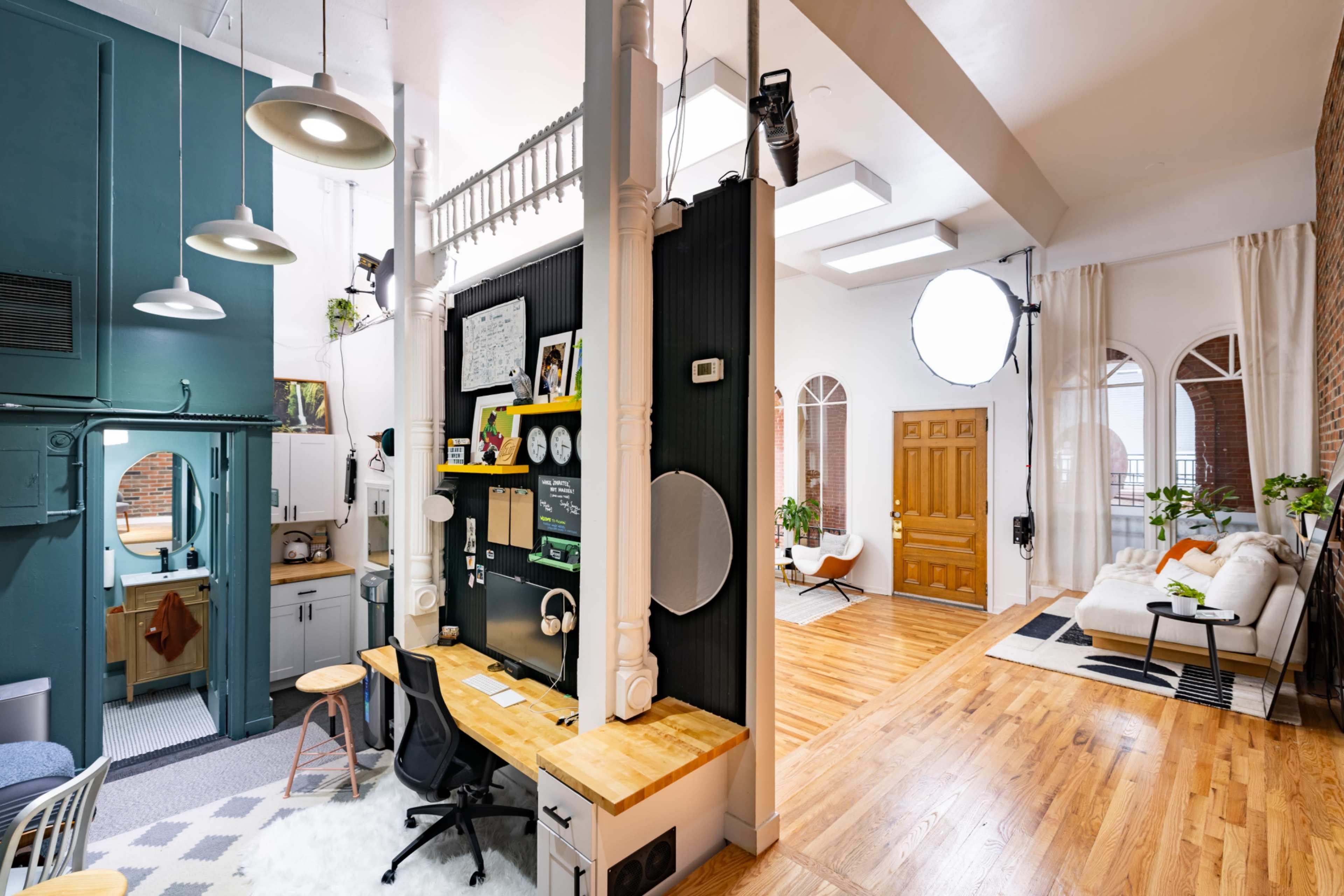 The image shows a spacious, modern interior featuring a work desk with a computer, a small kitchen area, and a cozy living room with large windows and a light-colored sofa.