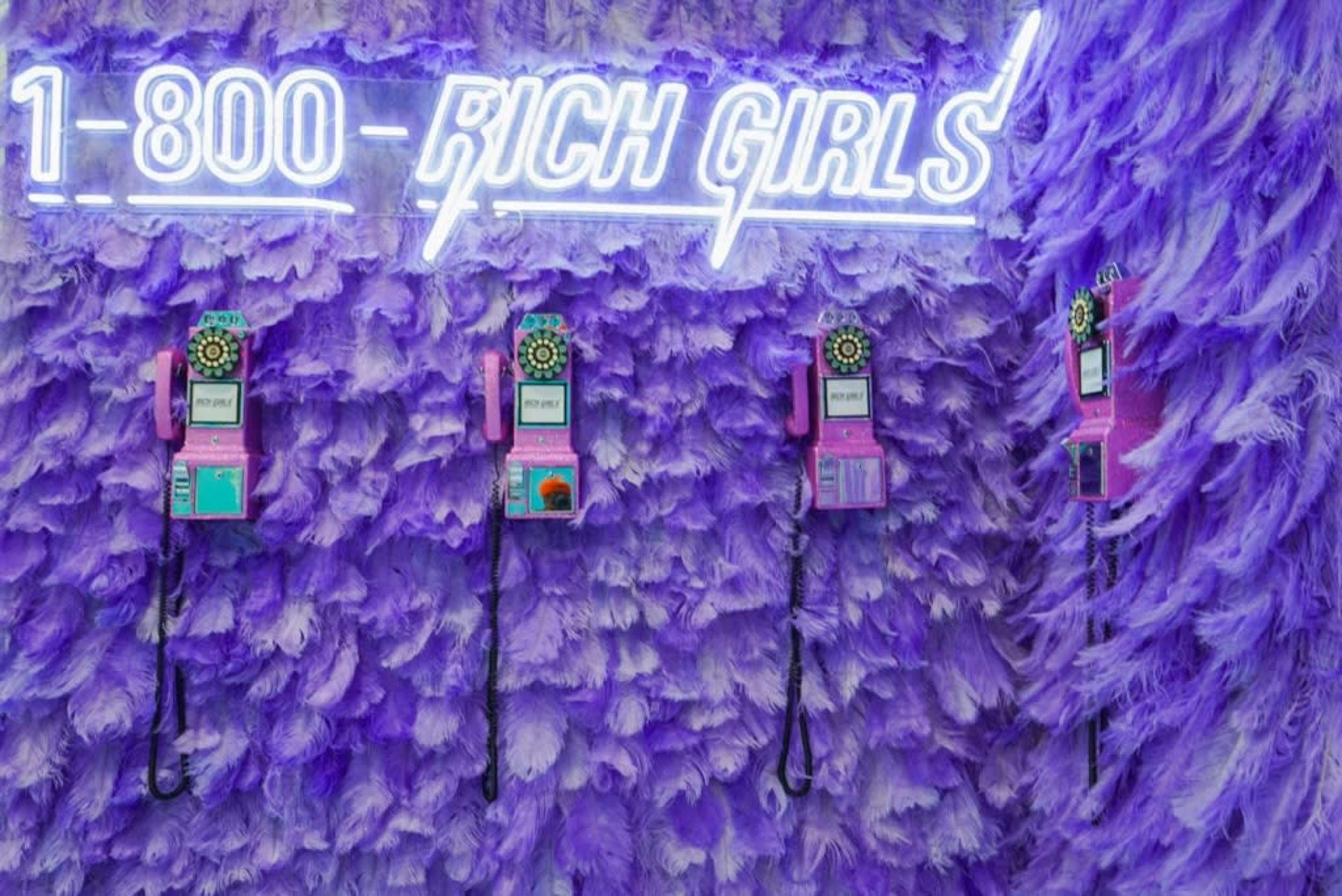 A wall covered in purple feathers with four vintage-style telephones hanging beneath a neon sign that reads "1-800-RICH-GIRLS."