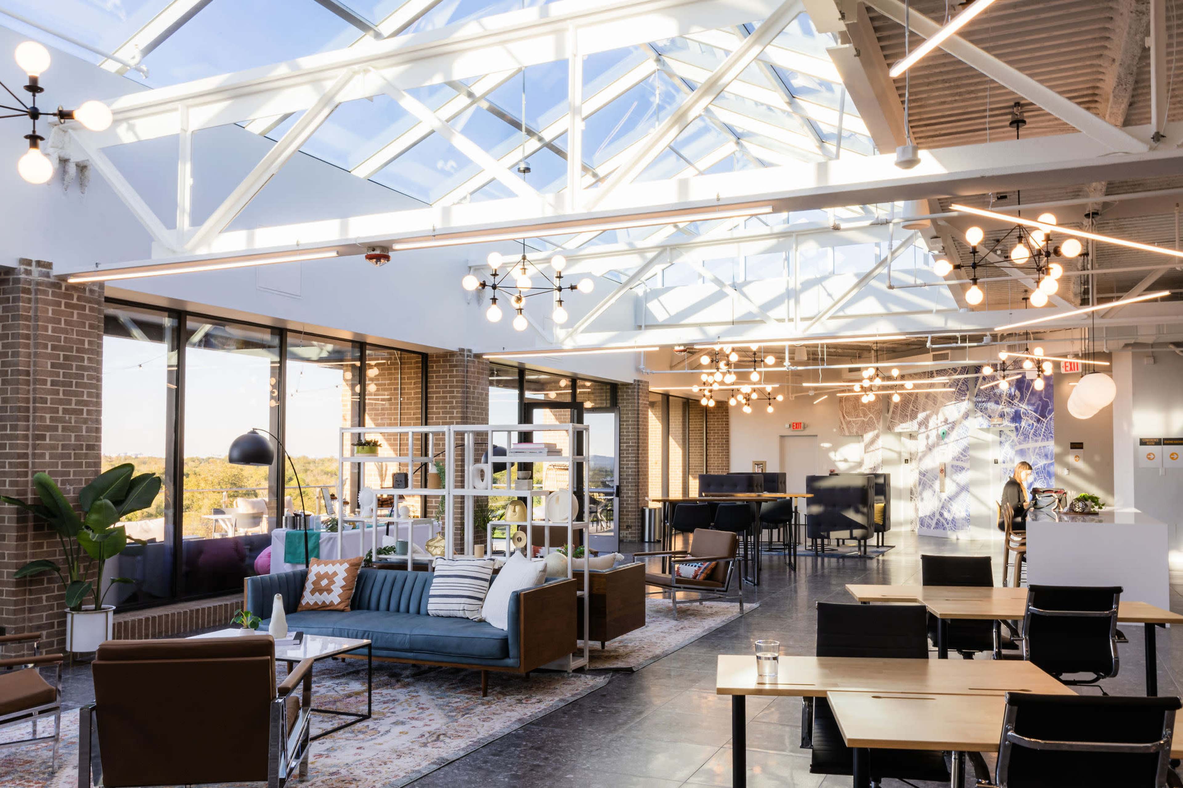 The image shows a modern, brightly lit open space with large windows, comfortable seating areas, and a mix of tables and lounge furniture.