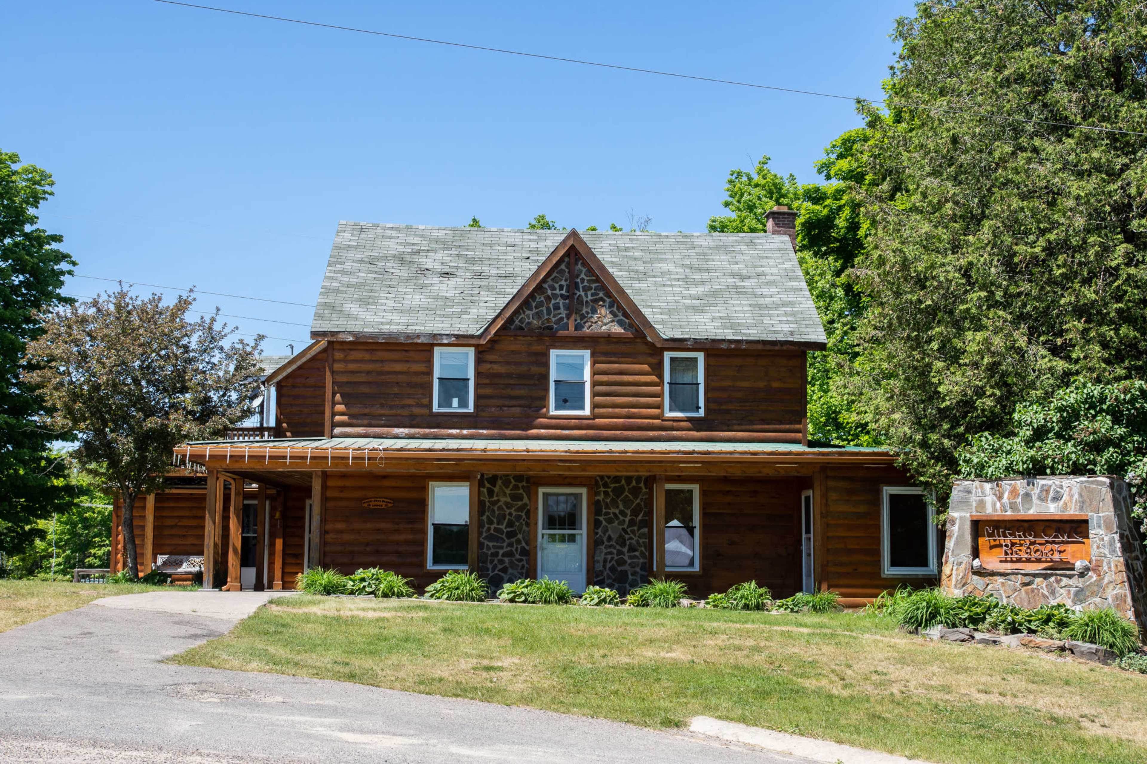 A two-story log cabin with a stone foundation and a sign, surrounded by greenery and trees.