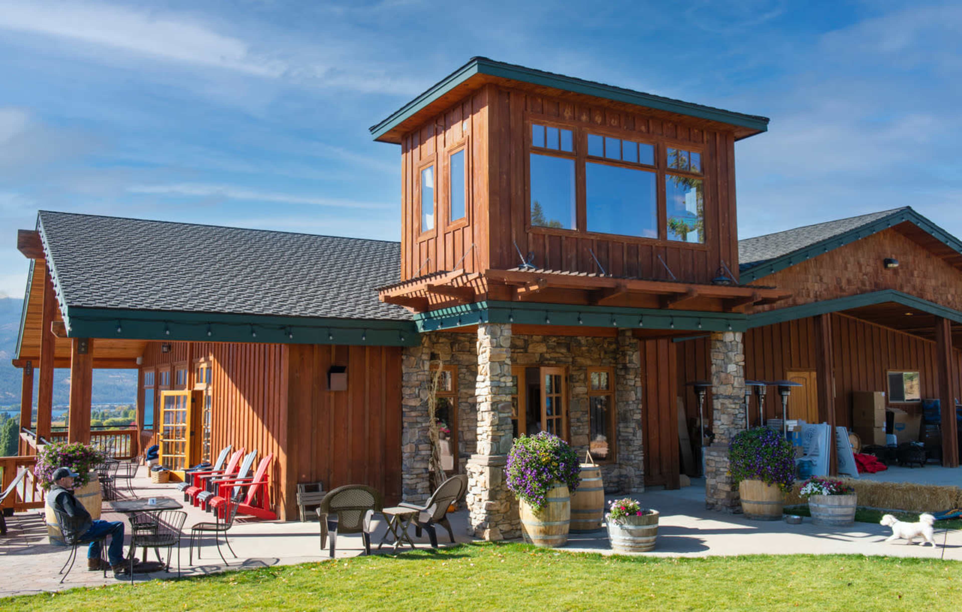A wooden building with large windows and a stone base, surrounded by outdoor seating and planters on a grassy lawn.