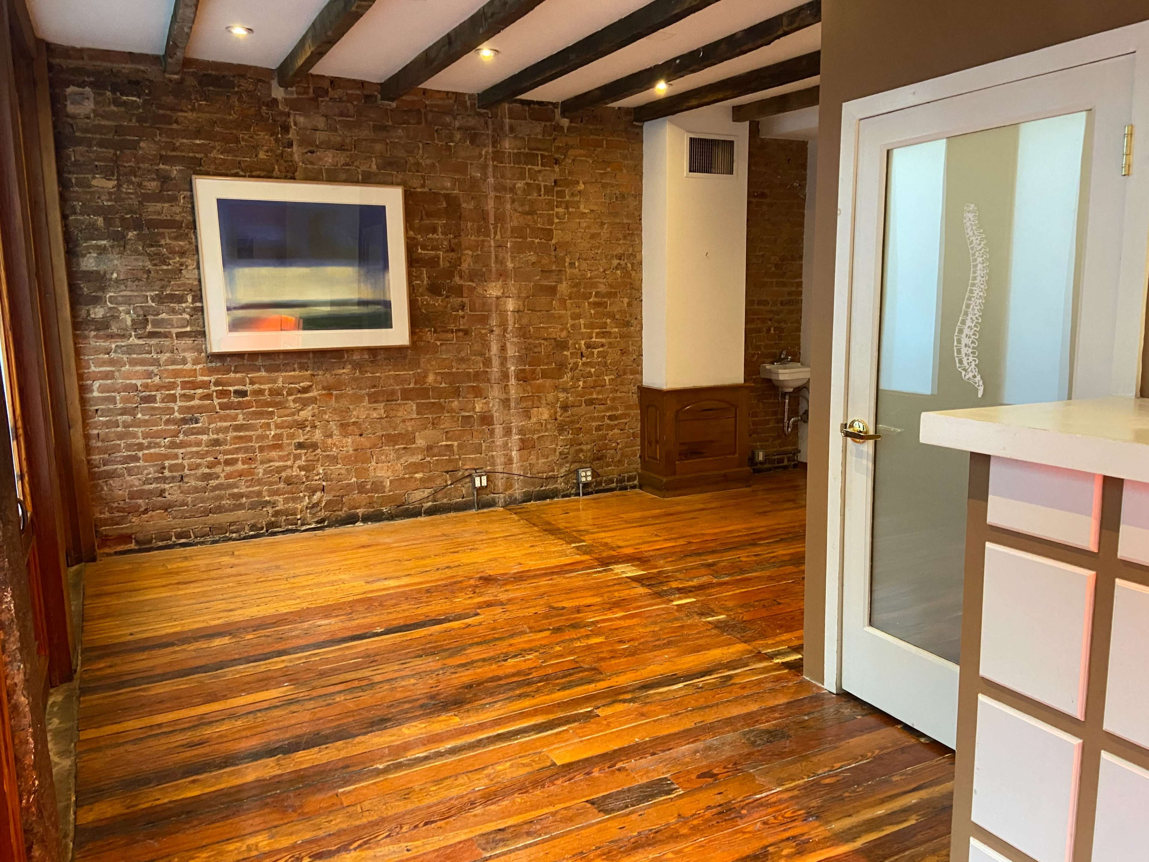An empty room with exposed brick walls, wooden flooring, and a framed artwork hanging on the wall, along with a doorway and a small sink visible in the background.