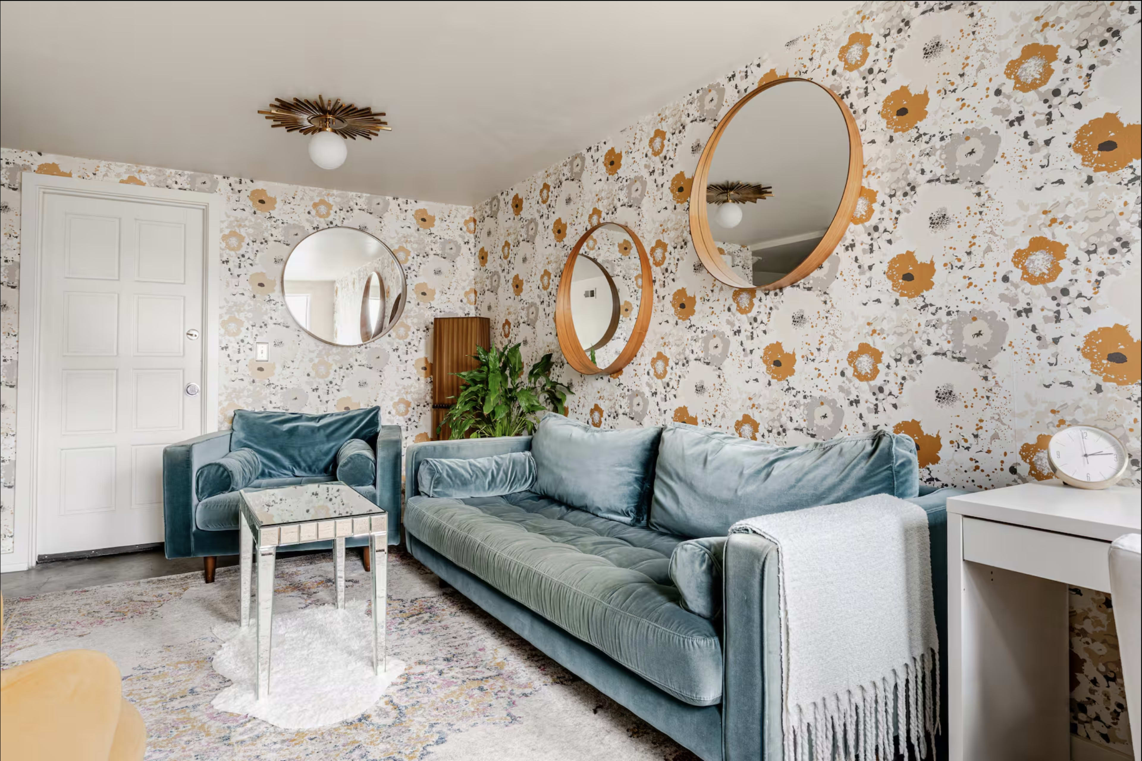 The image shows a living room with a teal velvet sofa, two teal armchairs, a decorative table, and three round mirrors on a floral-patterned wall.