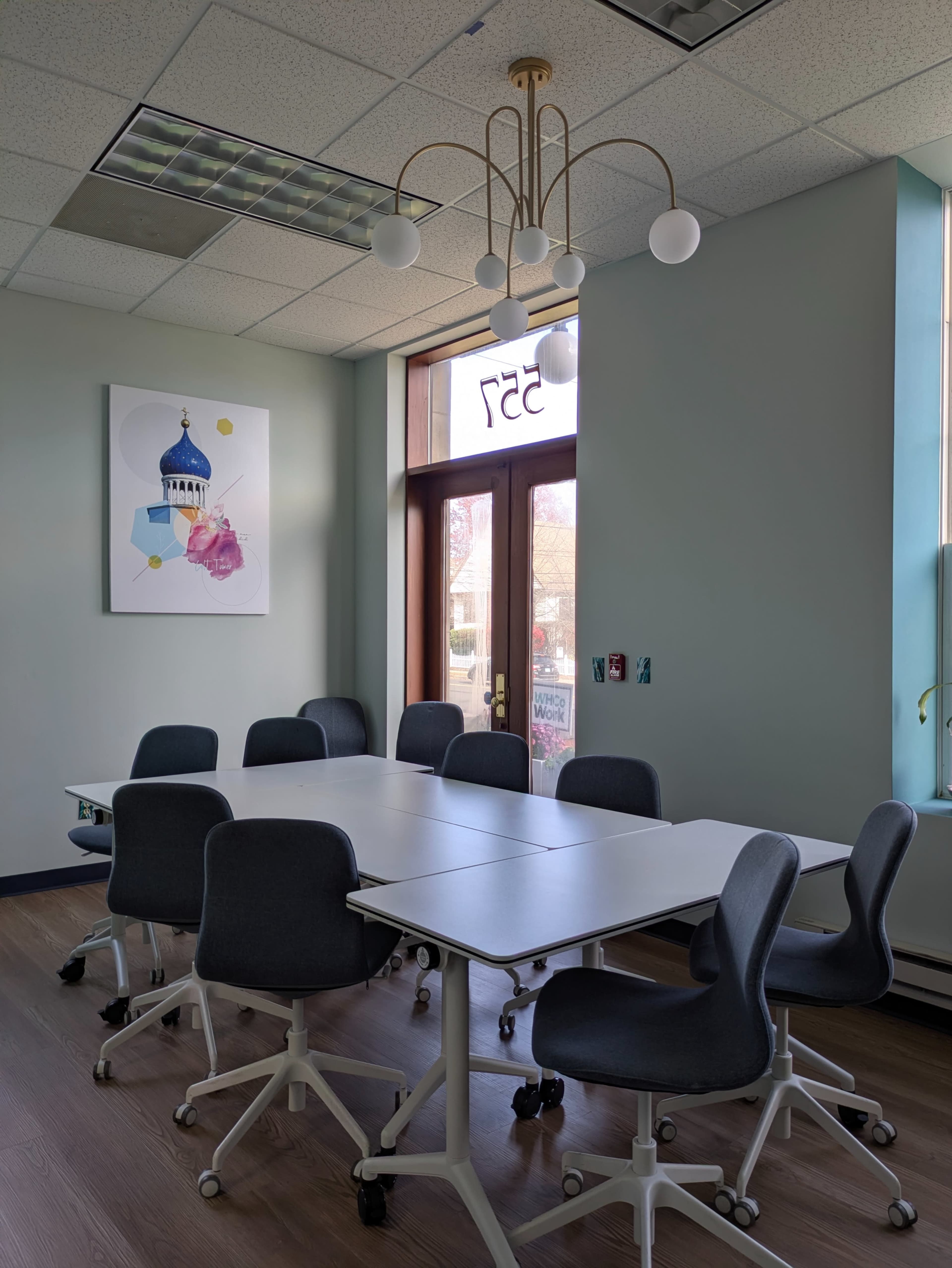 A bright conference room features a large white table surrounded by eight wheeled chairs, with a decorative chandelier above and a piece of artwork on the wall.