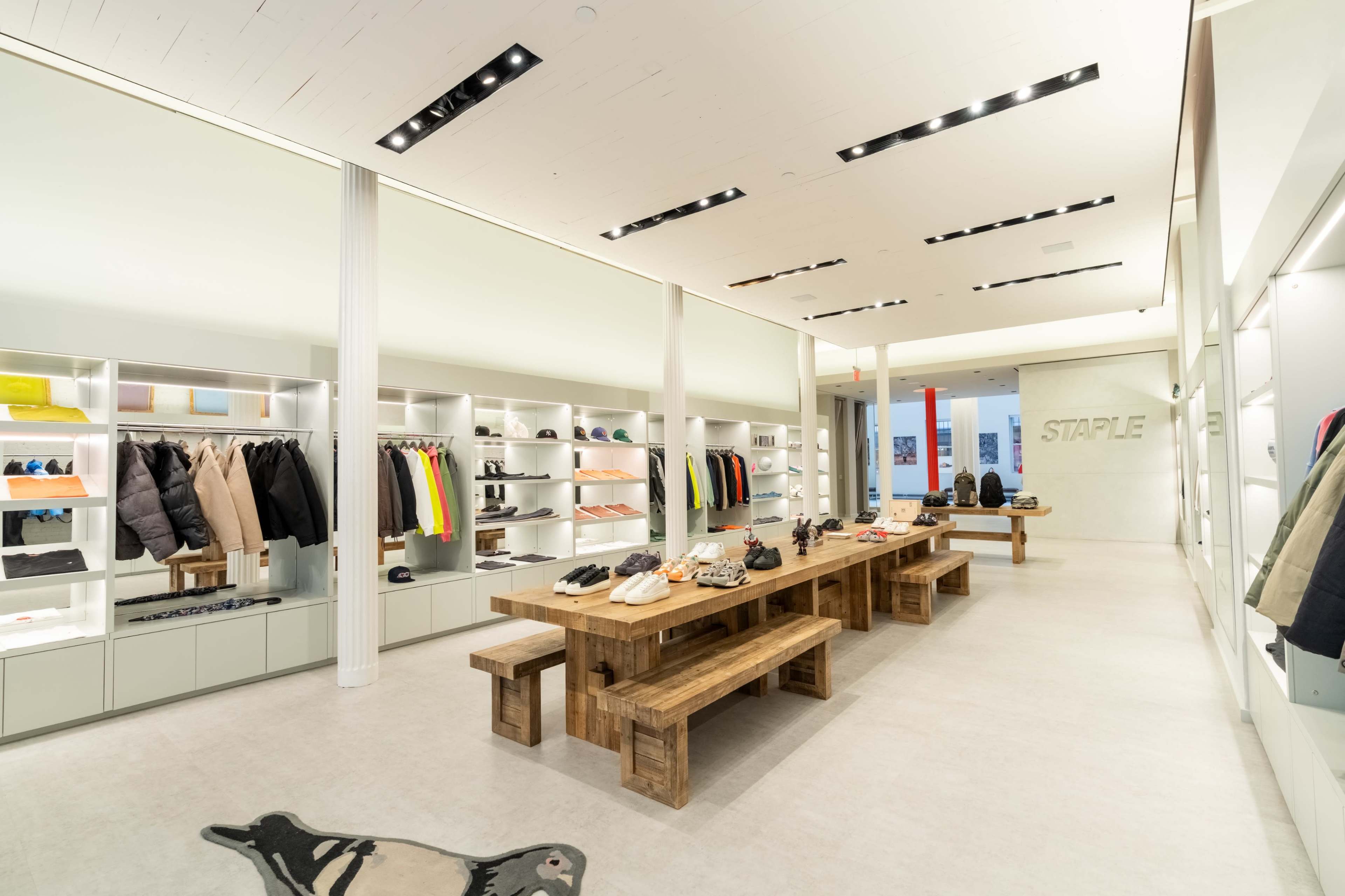 The image shows a modern retail store interior featuring neatly arranged clothing and shoes on display shelves, with a central wooden table surrounded by benches.