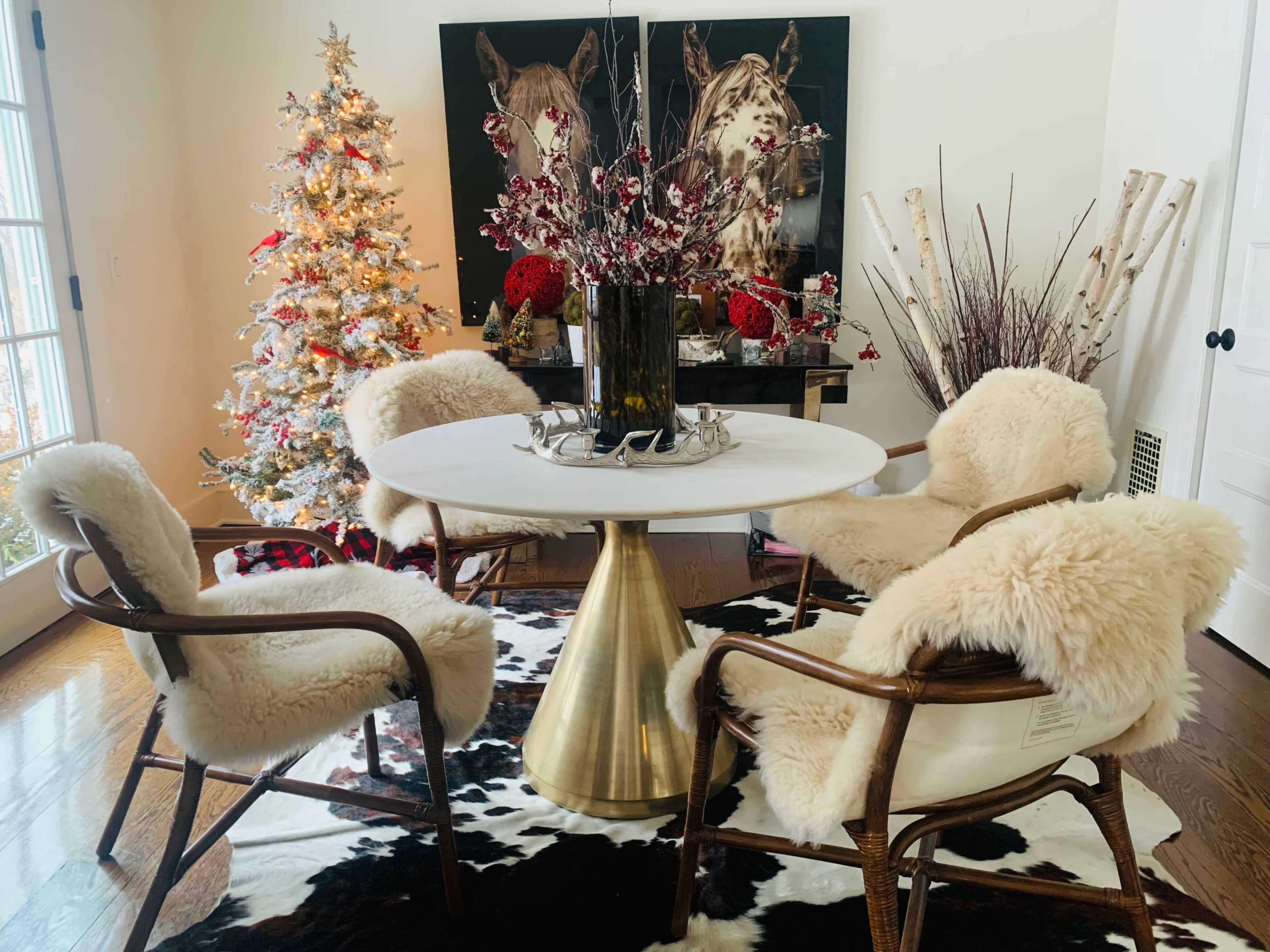 A round white dining table with four chairs, covered in faux fur, is set in a decorated room featuring a Christmas tree and artwork of horses.