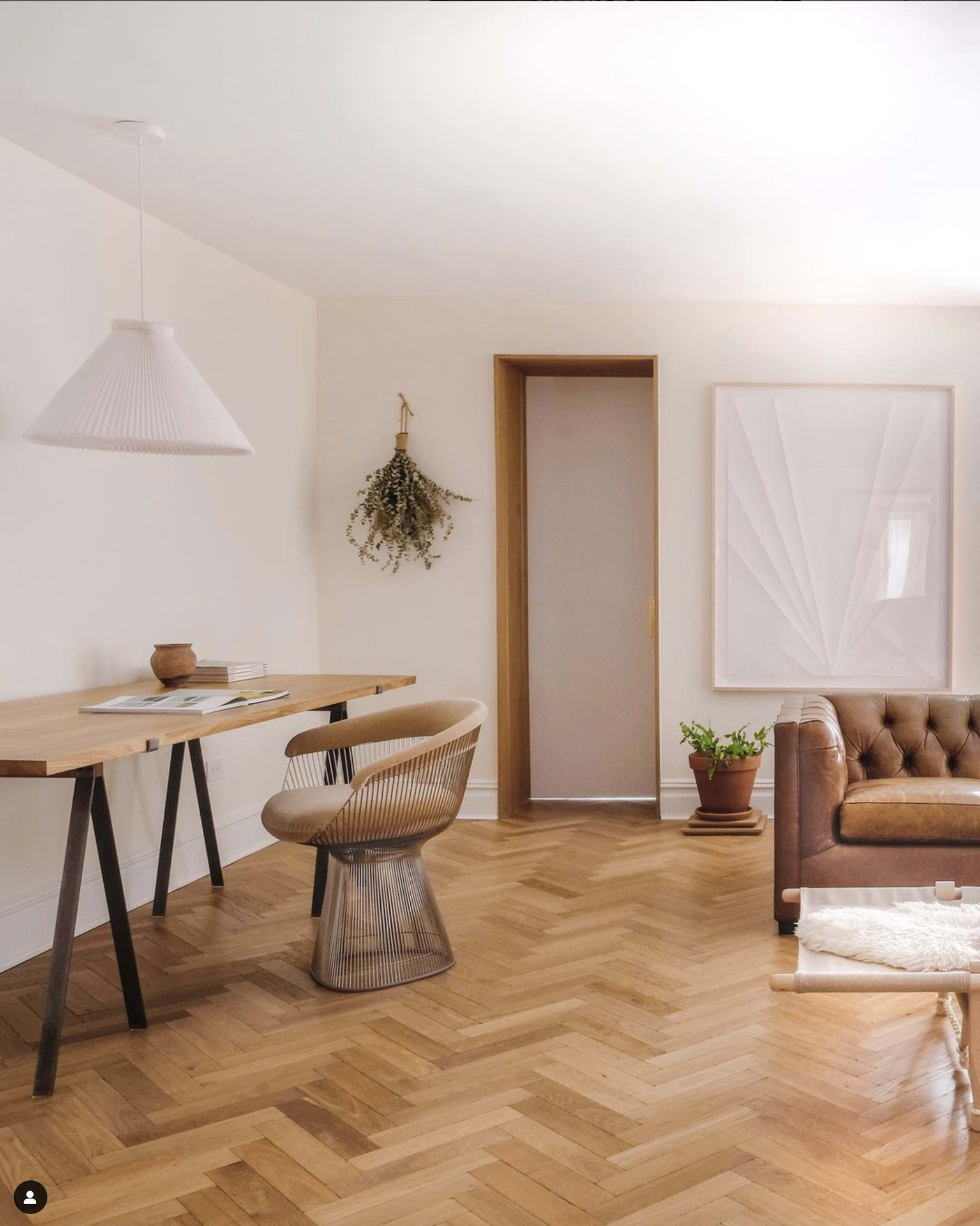 A modern living room features a wooden desk with a chair, a potted plant, and a leather sofa on herringbone-patterned flooring.
