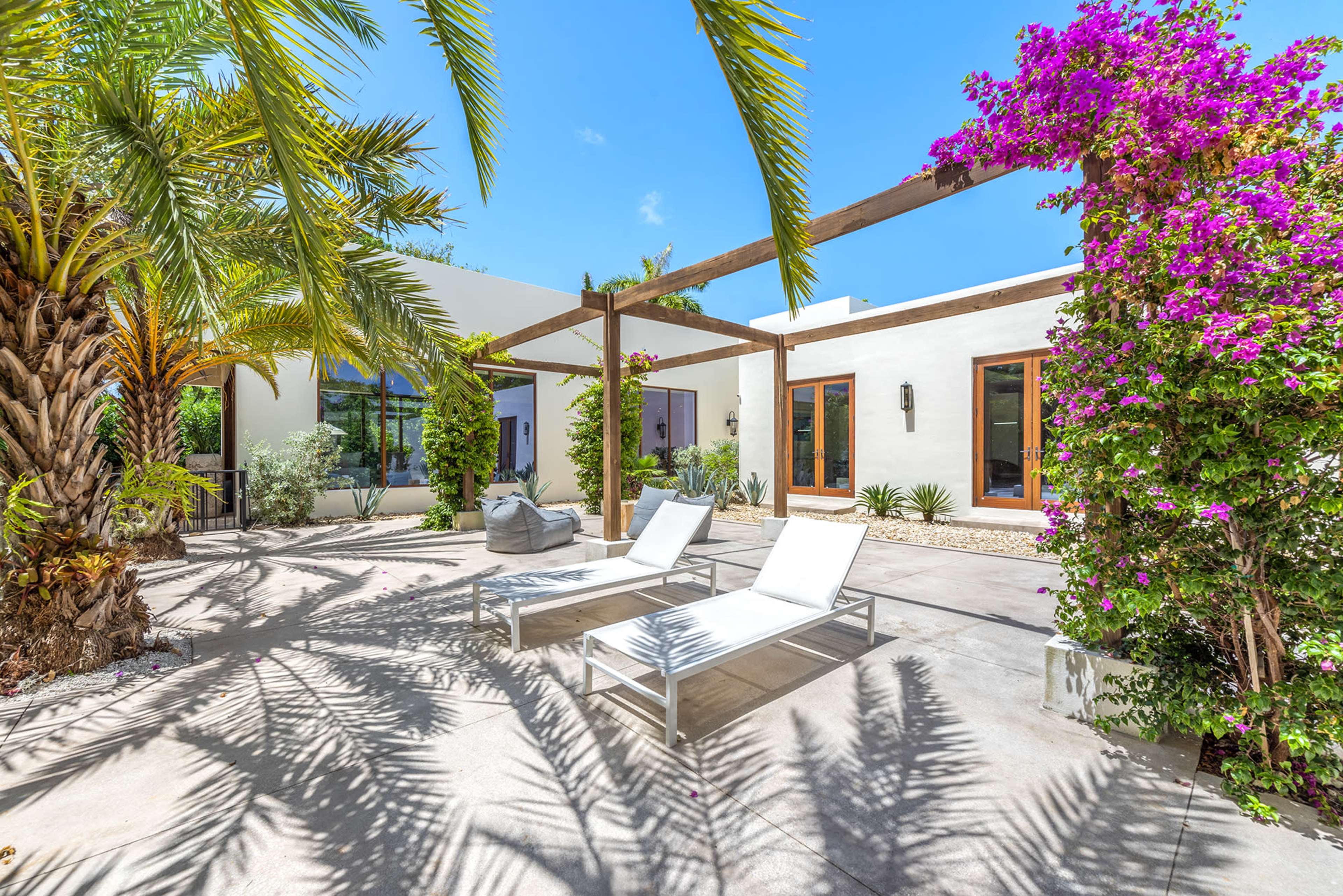 A patio area features white lounge chairs beside a palm tree and blooming bougainvillea under a clear blue sky.