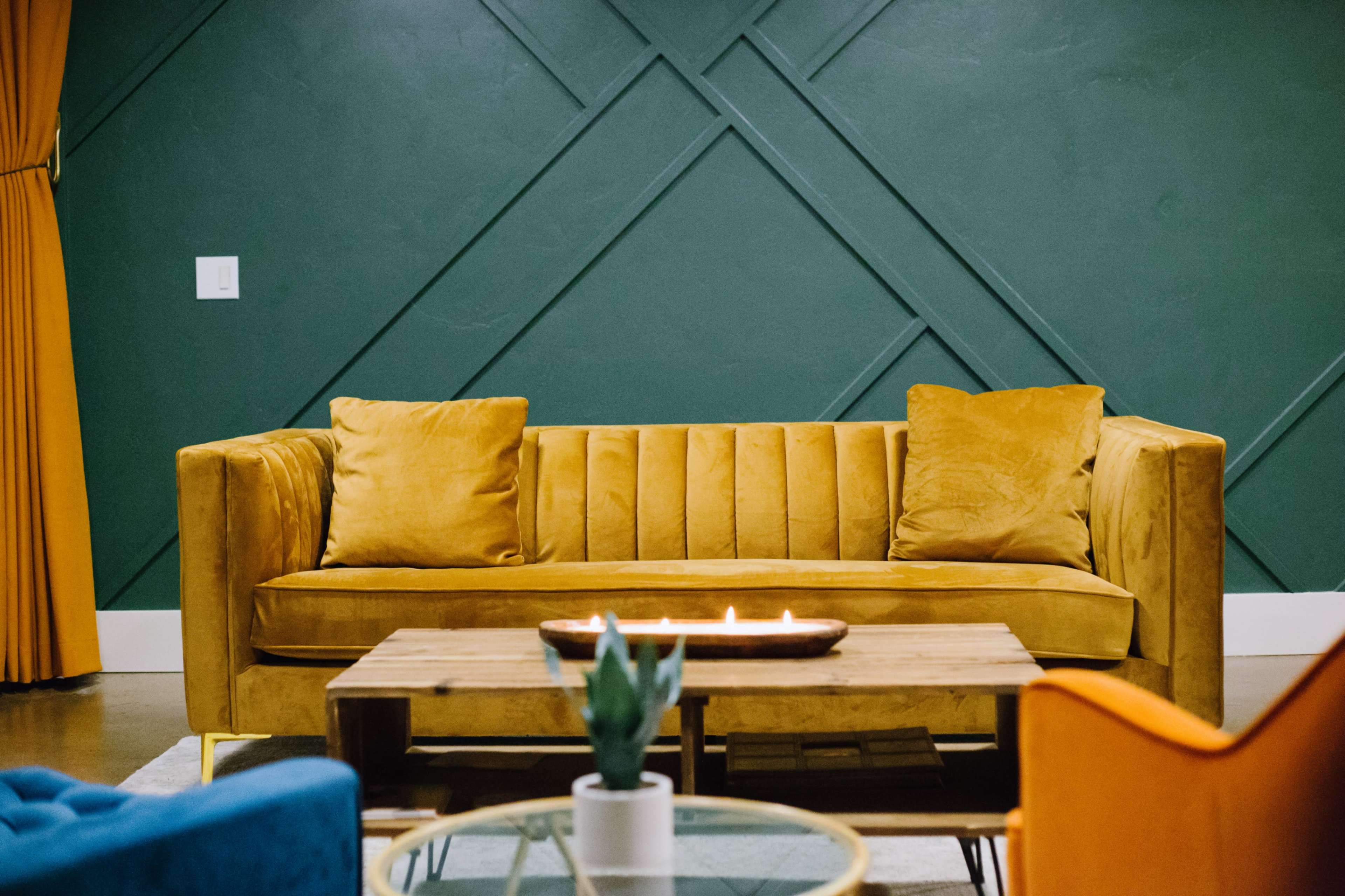 A yellow sofa with plush cushions, a wooden coffee table with candles, and a green accent wall with a geometric design.